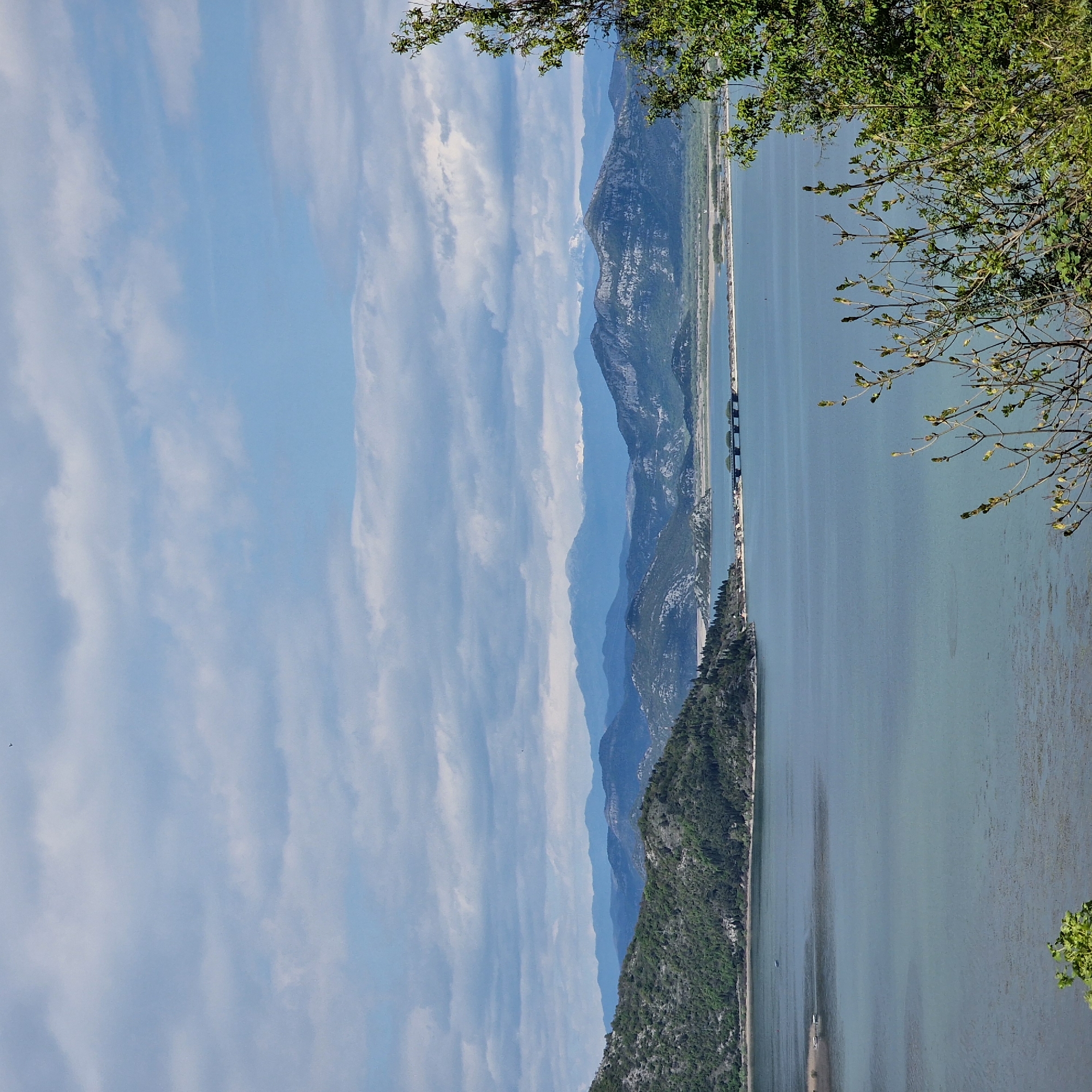 The image depicts a body of water (likely Skadar Lake) extending into the distance, flanked by mountains on the left and greenery in the foreground on the right. A causeway with a bridge segment stretches across the lake. The sky is partly cloudy with a blue hue. The location is likely Godinje, Montenegro, given the landscape characteristics.