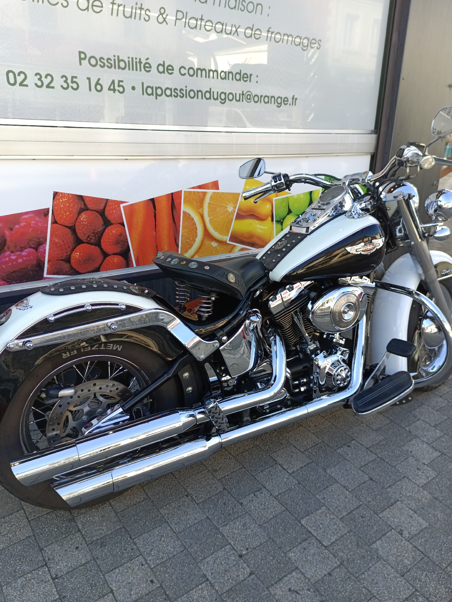 In Le Neubourg, France, a parked black and white motorcycle with chrome accents is captured in a medium, eye-level shot. The motorcycle is positioned on a cobblestone sidewalk, with its rear wheel and exhaust pipes prominently featured in the foreground. Behind the motorcycle, a white sign is visible, advertising fruits, platters, and cheeses, along with contact information including a phone number and an email address. The sign also displays colorful images of fruits and vegetables. The overall scene suggests a commercial setting, possibly outside a shop or market. No people are present in the image.