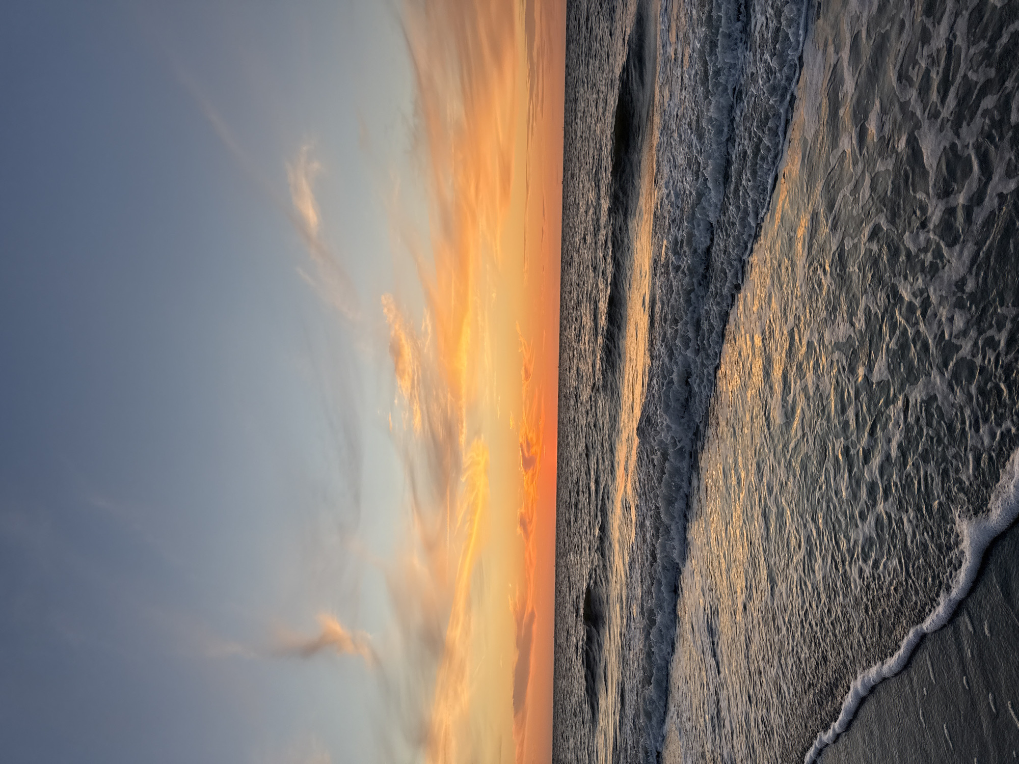 This is a vertical, eye-level, full shot of a beach at sunset. The ocean fills the bottom two-thirds of the frame, with waves rolling in towards the shore. The water is a dark blue-grey, reflecting the warm hues of the sunset. A thin line of white foam marks the edge of the water on the dark sand in the foreground. The sky above is a gradient of soft blues at the top, transitioning to vibrant oranges and yellows near the horizon. Wispy clouds are scattered across the sky, catching the last light of the day. The overall impression is one of tranquility and natural beauty.