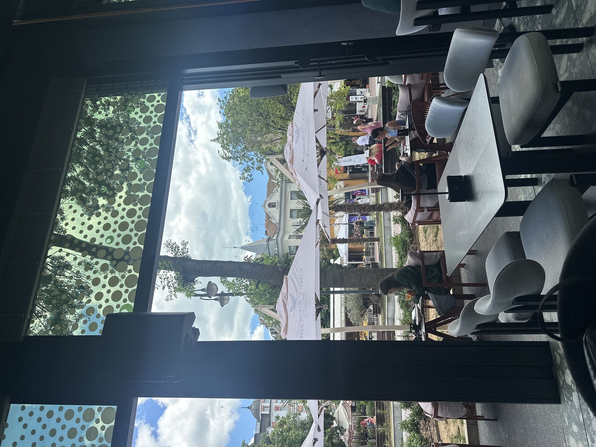 The image captures an outdoor seating area of a cafe or restaurant, viewed through a large window. In the foreground, several dark-legged tables with light-colored chairs are visible. Through the window, an outdoor scene unfolds under a cloudy sky. Several white umbrellas are set up, some displaying branding. People are seated at tables outside, some engaged in conversation. In the background, there are trees, lampposts, and buildings with architectural details typical of a European city. One building features a distinct domed roof, and another has a pointed spire. The overall atmosphere suggests a leisurely day in a public square or park.