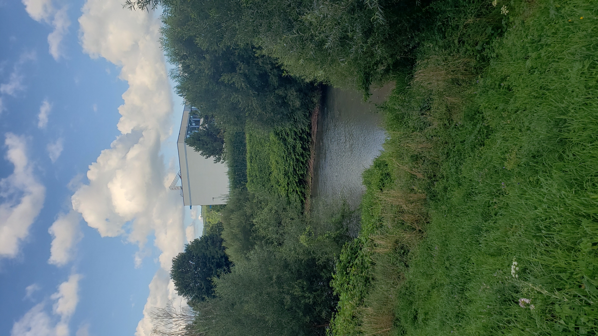 The image depicts a natural scene with a body of water in the midground, bordered by lush green vegetation. In the background, a modern white building with blue-tinted windows is visible, surrounded by trees and a neatly trimmed hedge. The sky above is blue with scattered white clouds, indicating fair weather. The foreground consists of a grassy slope. Without further information or visual cues within the image, it is impossible to definitively place this scene in the city of Prešov, Slovakia, or any specific city.