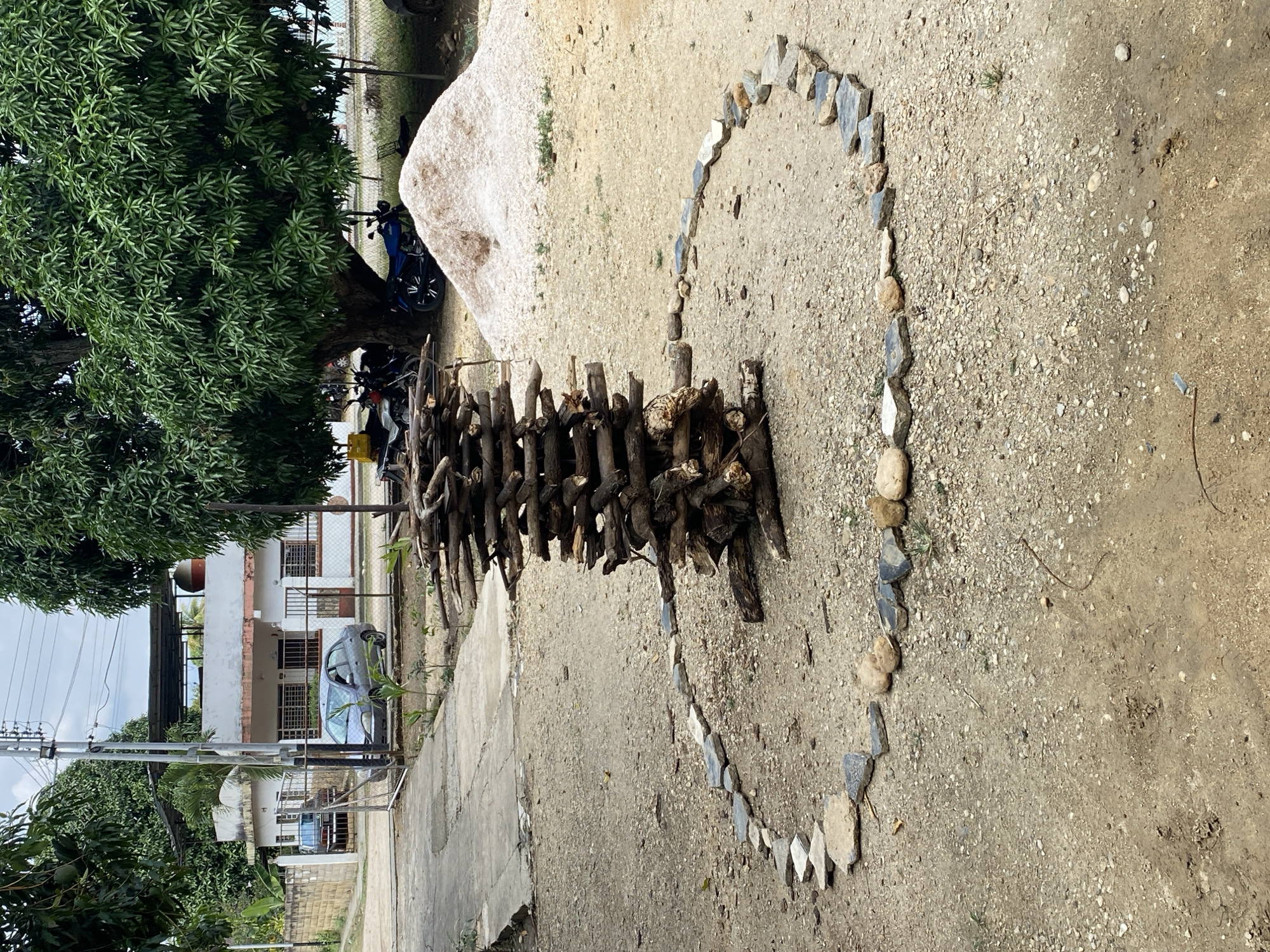 The image depicts an outdoor scene in what appears to be Tinaquillo, Venezuela. On a dirt and concrete surface, a heart shape is formed by rocks, and a collection of sticks is placed within the heart. In the background, there are trees, a large boulder, a building, vehicles, and utility poles. The scene suggests a possibly artistic or decorative arrangement of natural materials.