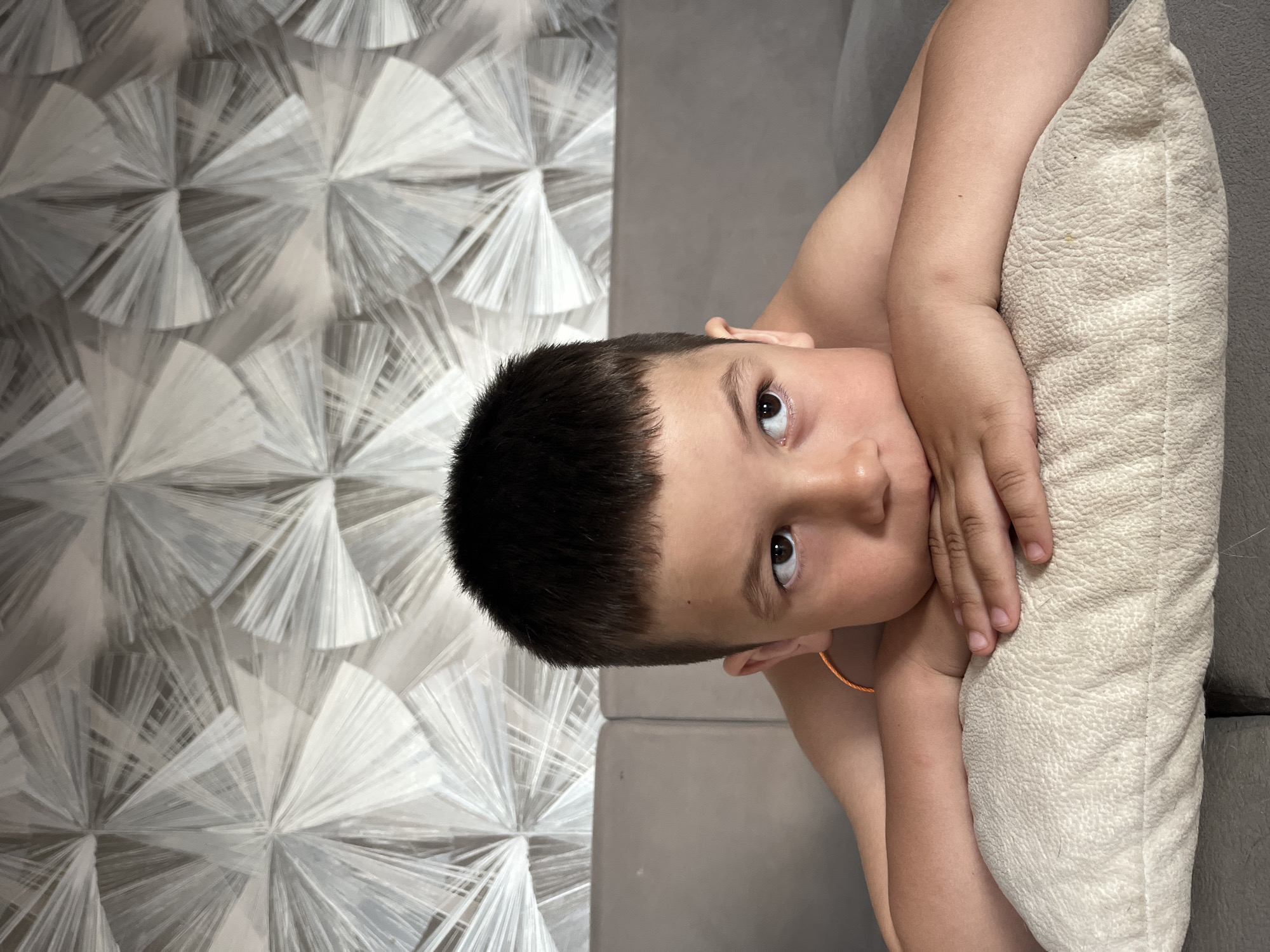 A young child is reclining on a gray sofa with a light beige textured pillow. The child has dark hair and is looking up at the camera. The child's arms are crossed, with hands near their face. The background consists of a wall with a geometric fan-like pattern. The location is unspecified.