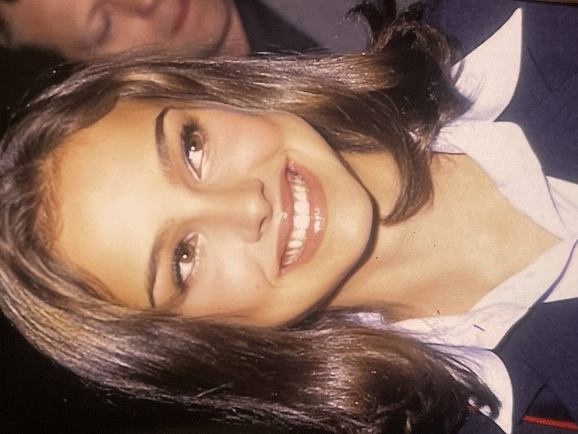 A close-up portrait of a young woman, tilted diagonally, fills the frame. She has warm-toned skin, dark brown hair, and bright brown eyes. A wide, joyful smile reveals her full set of white, even teeth, enhanced by glossy lips. Her eyebrows are dark and well-defined. She is dressed in a white collared shirt, worn under a dark navy jacket or blazer. Behind her and slightly above, a blurred portion of another person's face is visible, suggesting a social environment. The background is dark and out of focus, indicating an indoor setting or an evening event. The overall mood conveyed is happy and positive. No text or specific location cues relating to Poltava, Ukraine are visible in the image.