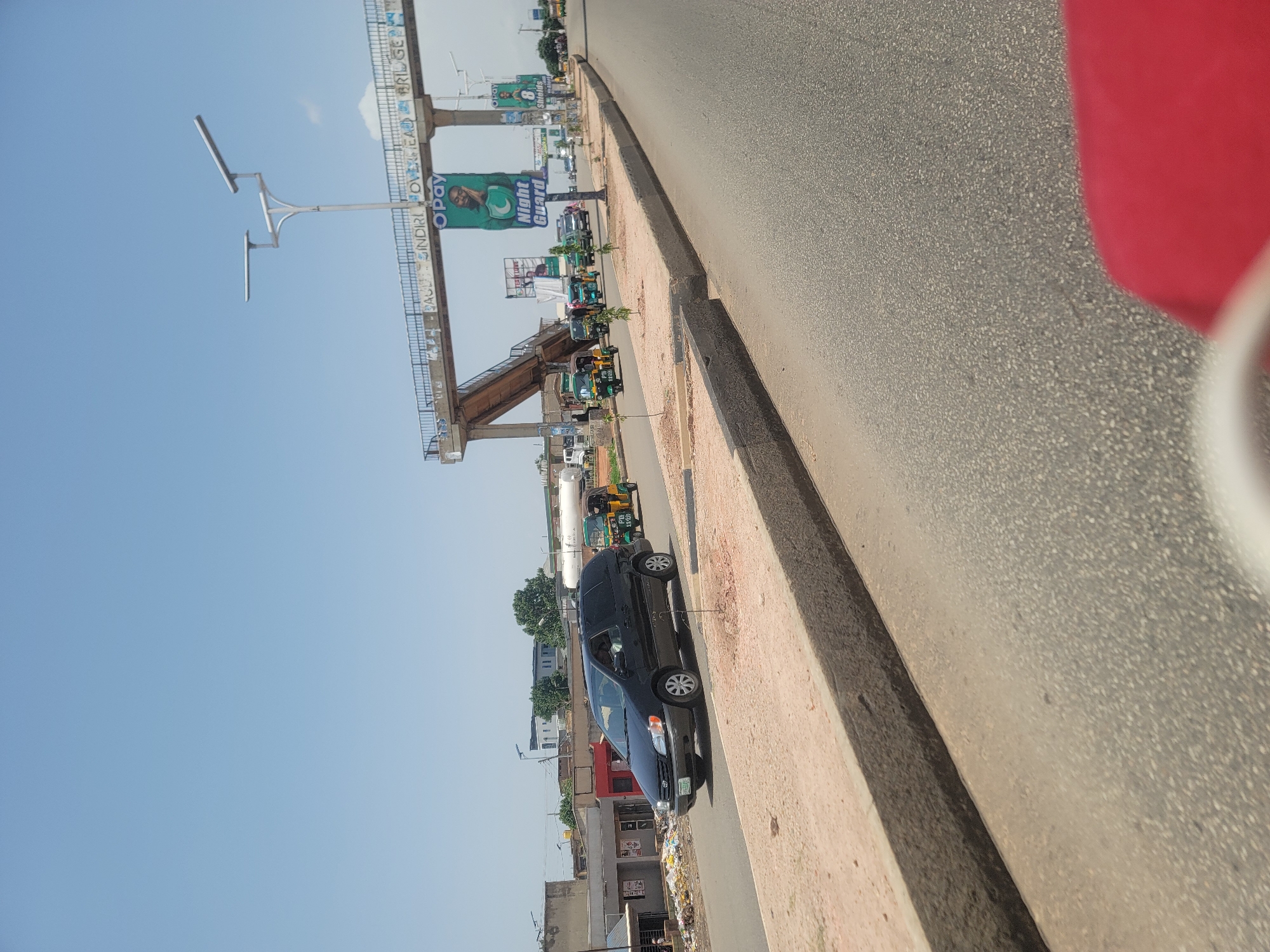 This media file shows a street scene in Gura Top, Nigeria, on a clear day. The perspective is from a moving vehicle, looking out at the roadside. An overhead bridge structure dominates the background, with various advertisements displayed on its sides. One prominent advertisement features the OPay logo and a portrait of a man with the text "Night Guard".
Along the roadside, there is a row of buildings and a pedestrian walkway. Several vehicles are visible, including a black SUV parked by the side of the road and several three-wheeled vehicles, commonly known as Keke Napep, which appear to be moving or parked. There are also some trees and streetlights. The overall impression is that of a bustling urban environment, likely during daylight hours given the bright conditions and clear sky. The pavement and roadside verge are visible, with some debris scattered on the ground. The presence of the overhead bridge and the types of vehicles suggest a developed area within the city.