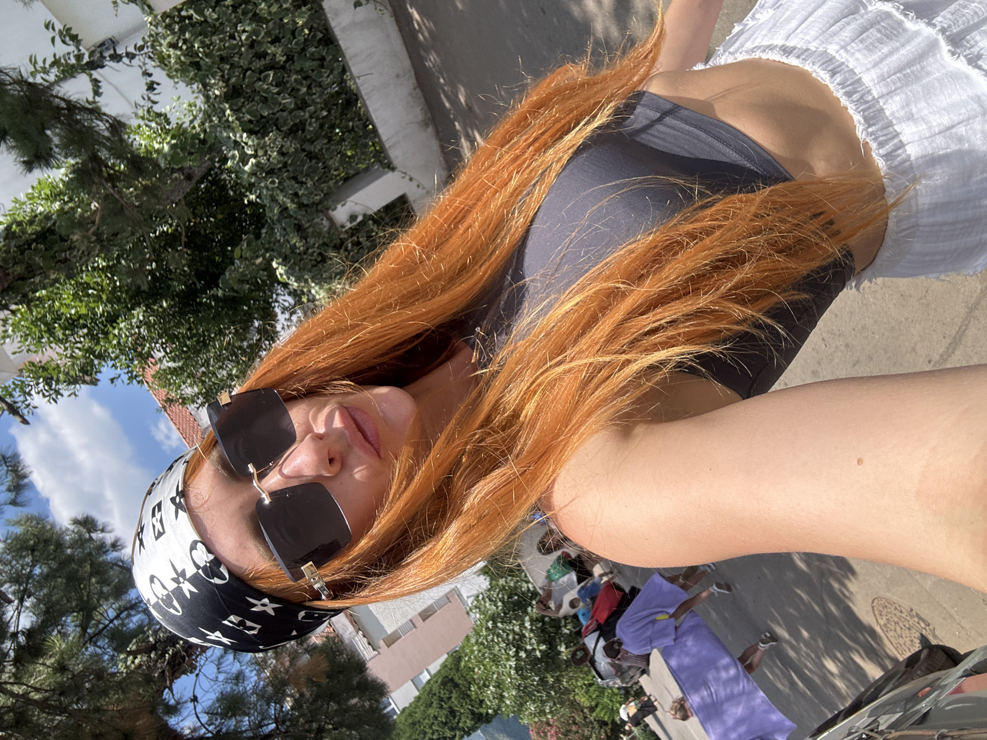 A woman with bright orange hair, wearing a black and white bandana and large black sunglasses, takes a selfie outdoors. She is wearing a dark gray crop top and a white ruffled skirt. The background shows a street scene in Budva, Montenegro, with trees, buildings, and several other people walking. One person in the background is carrying a long, purple object. The sun is shining, casting shadows on the ground.