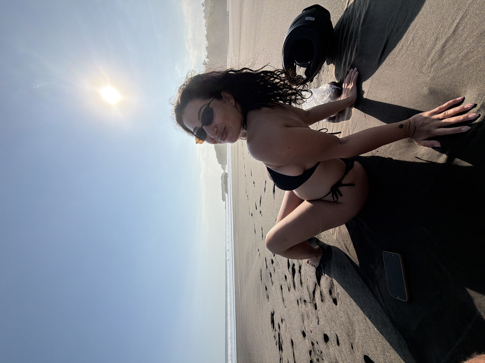 The image captures a woman seated on a black sand beach in Selemadeg, Indonesia, under a clear, light blue sky with a bright sun causing lens flare. The woman, with dark curly hair and dark sunglasses, wears a black two-piece bikini and smiles towards the viewer. She rests on a dark mat or towel, leaning on her right arm with her left hand on the sand, her legs bent. A small, dark tattoo is visible on her right wrist. To her right lies a black motorcycle helmet with "DOT" markings and a clear plastic bag containing unspecified items. A dark smartphone with a light-colored case rests on the mat to her left. The background features the ocean with white breaking waves, distant land formations, and tropical vegetation, all illuminated by strong sunlight that casts long shadows across the dark, footprint-laden sand.