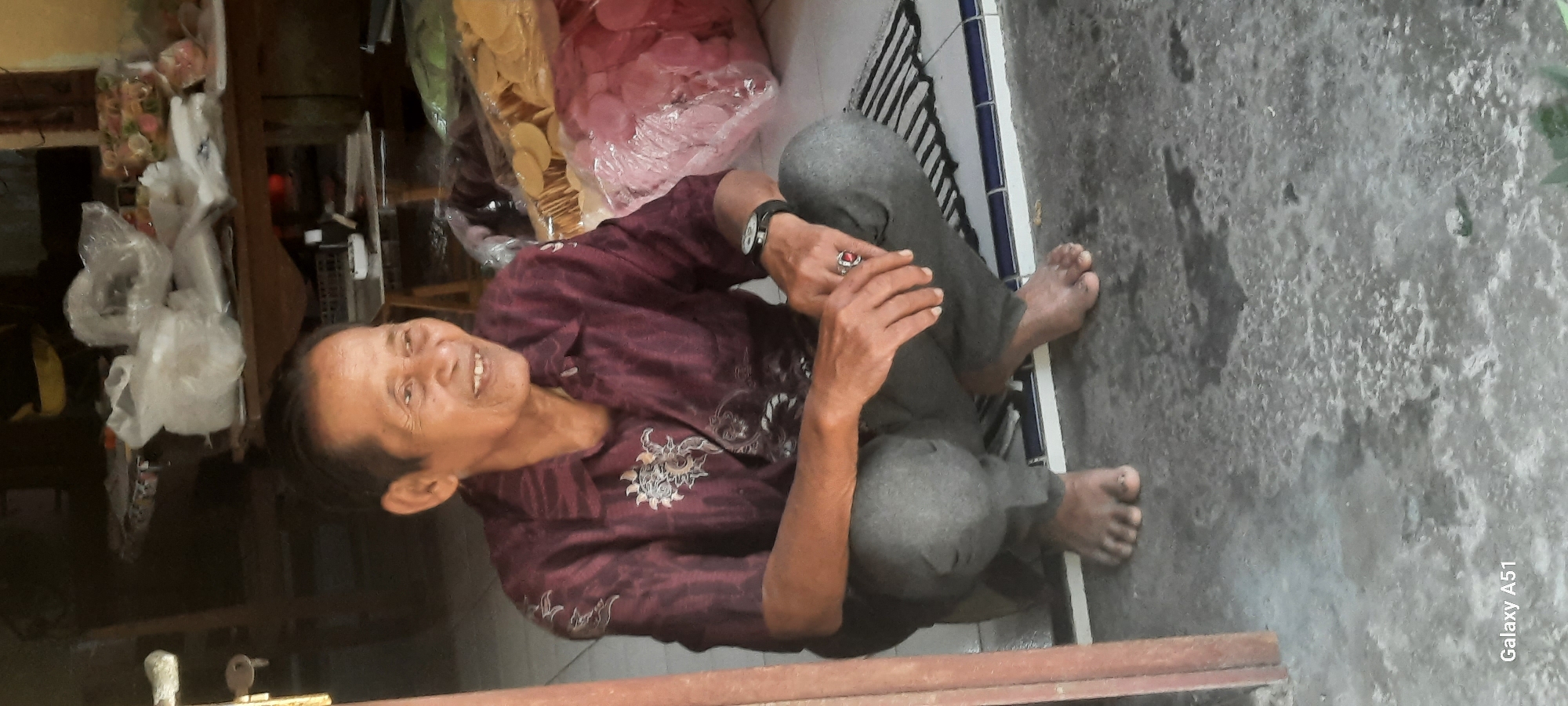 The image shows an elderly East Asian man squatting on a tiled floor in what appears to be an open-air market or shop in Ploso, Indonesia. The man is wearing a dark red patterned shirt and dark grey pants. His hands are clasped in front of him, and he is wearing a ring with a red stone on his right hand. Large bags of what appear to be crackers or snacks are visible in the background, suggesting a commercial setting. The floor is a mix of tiled and concrete surfaces, with some water stains visible. The lighting suggests it is daytime, possibly with overhead sun. The overall impression is of a relaxed, casual scene within a busy commercial environment.