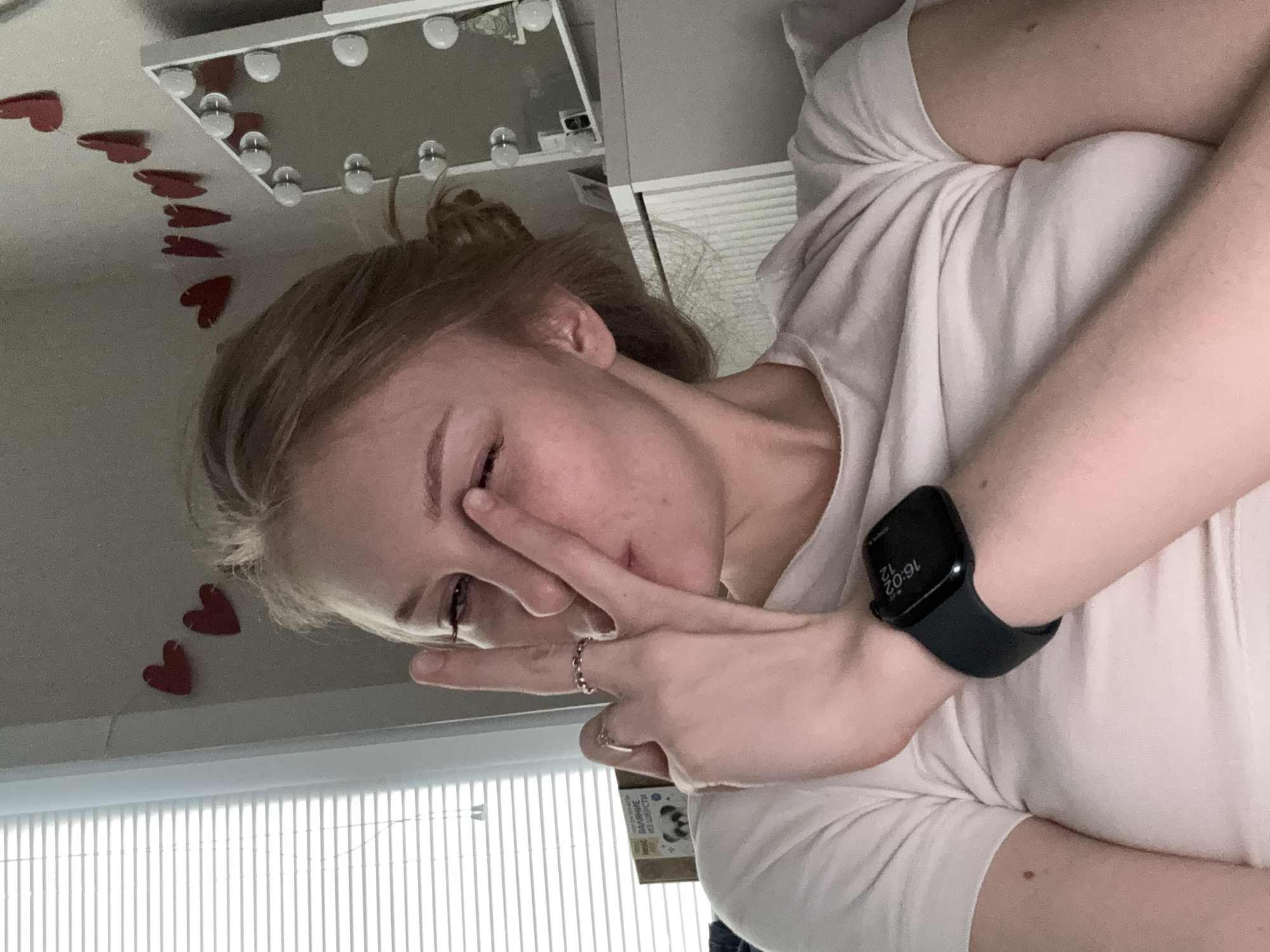 In the city of Perm, Russia, a young woman with blonde hair pulled into a bun is seen from the chest up, positioned indoors. She is wearing a light-colored t-shirt and has her right hand raised, with her index and middle fingers extended in a peace sign gesture, touching her face. A silver ring is visible on her left ring finger. She is also wearing a dark smartwatch on her left wrist. In the background, a white dresser with a mirror that has multiple light bulbs is visible. Red heart decorations are affixed to the wall above the dresser. To the left of the woman, a window with white horizontal blinds is partially visible. A box with text and an image of a panda is on a shelf to the left. The lighting suggests an indoor setting, possibly a bedroom, with a soft, ambient light.