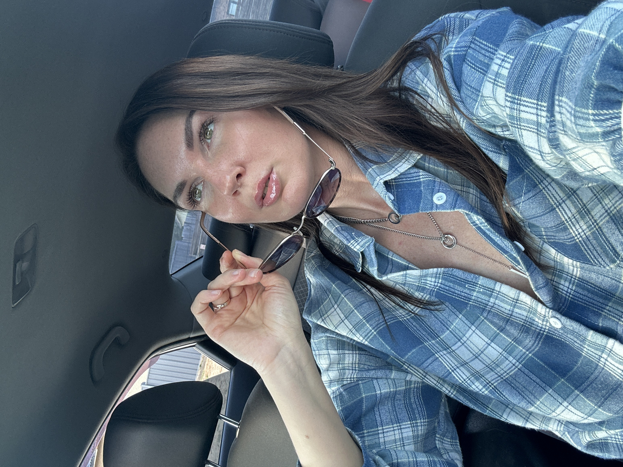 A woman is in the front seat of a car. She is wearing a blue and white plaid shirt. She is holding a pair of sunglasses in her right hand. She has long brown hair. She is wearing a silver necklace. The location of the photograph is unspecified, so this cannot be determined.