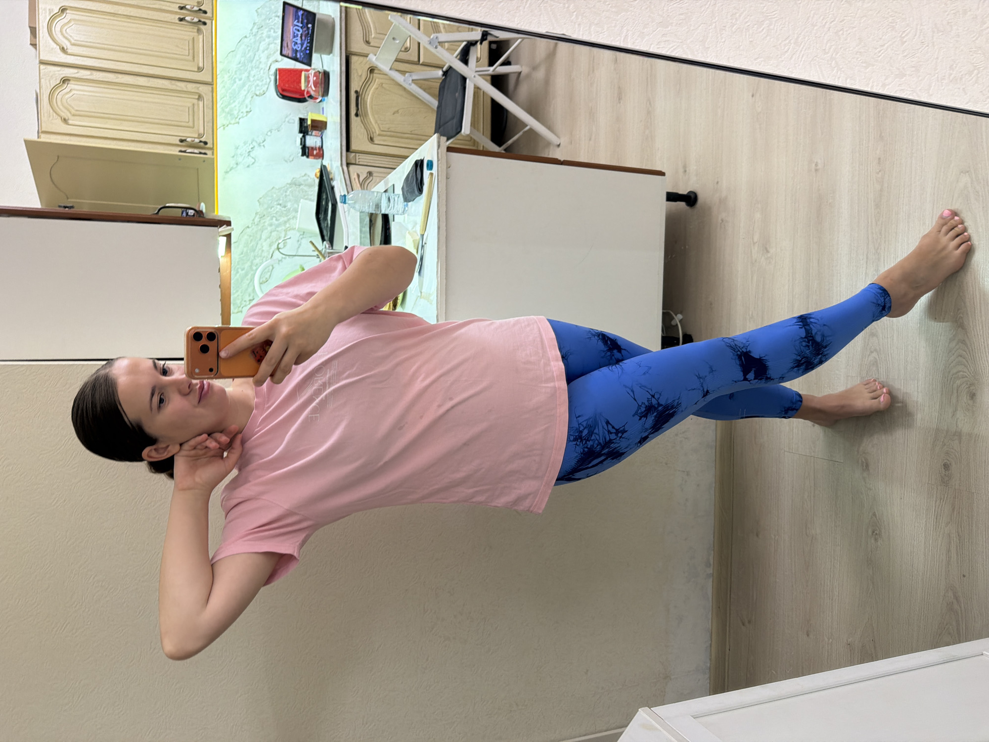 A woman is taking a selfie in front of a mirror. She is wearing a pink t-shirt and blue leggings. The background is a kitchen. The location is possibly Sochi, Russia.