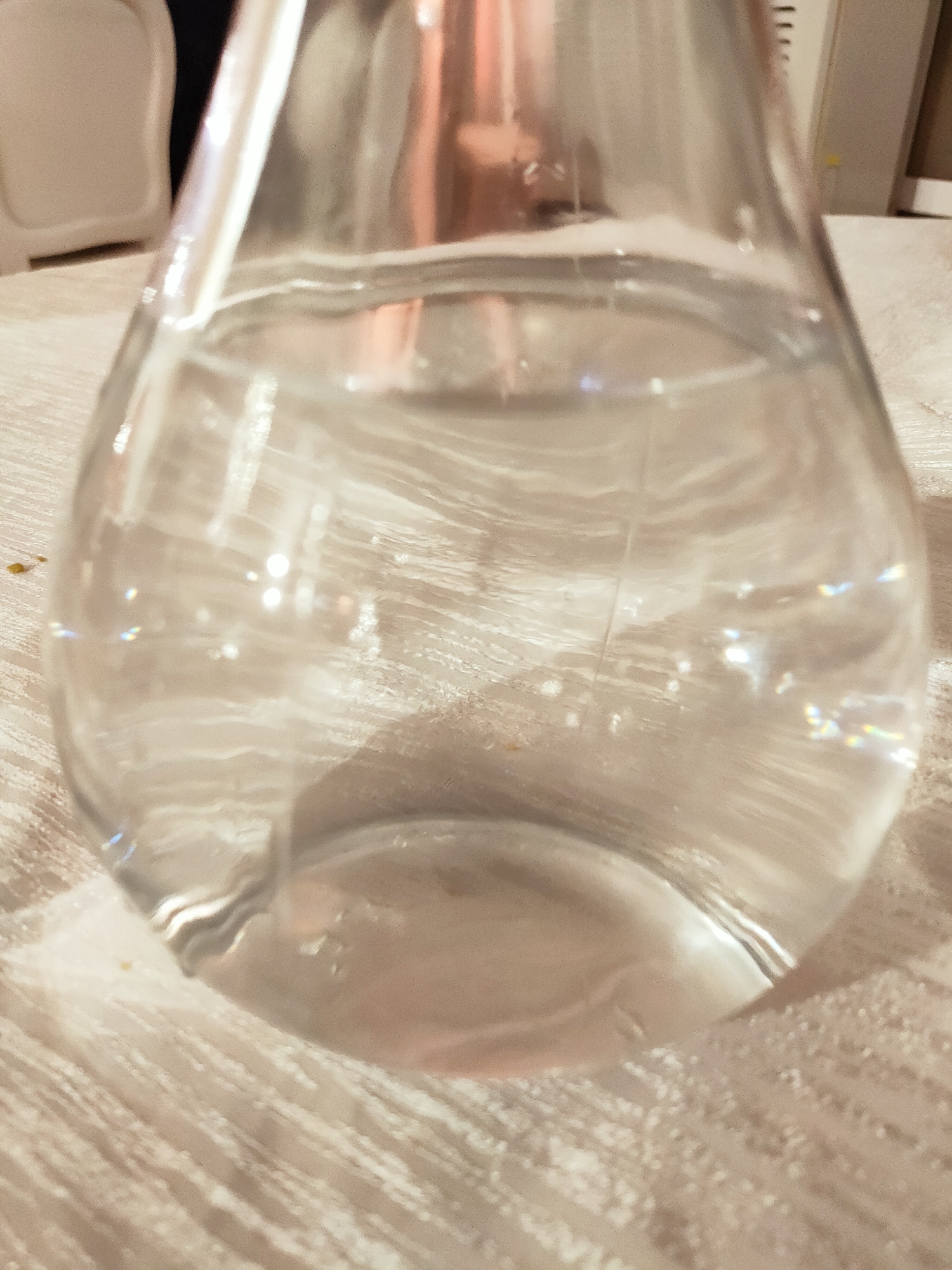 The image shows a close-up, low-angle shot of a glass carafe filled with clear liquid, possibly water. The carafe is resting on a textured, light-colored surface, which appears to be a tablecloth or placemat. The glass exhibits reflections and refractions of light, creating bright spots and rainbow-like prismatic effects. In the background, out of focus, a white chair or stool is partially visible on the left, and a white appliance with horizontal lines, possibly a refrigerator or a cabinet, is seen on the right. The overall lighting is dim and warm. There are no people visible in the image. The scene is indoors, on a table setting. The location in the city of Mornag, Tunisia is not discernible from the visual information provided in the image.