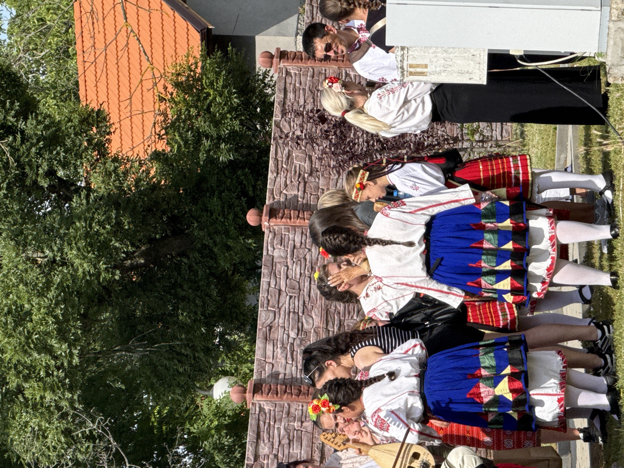 In the city of Pordim, Bulgaria, a group of people are gathered outdoors, dressed in traditional Bulgarian folk costumes. The costumes feature white embroidered blouses and colorful skirts with geometric patterns. Some individuals are wearing flower headbands. A person on the left is holding a stringed instrument, possibly a kaval or gudulka. The background includes a stone wall, lush green trees, and a house with a red tiled roof. The atmosphere appears festive, suggesting a cultural event or celebration.