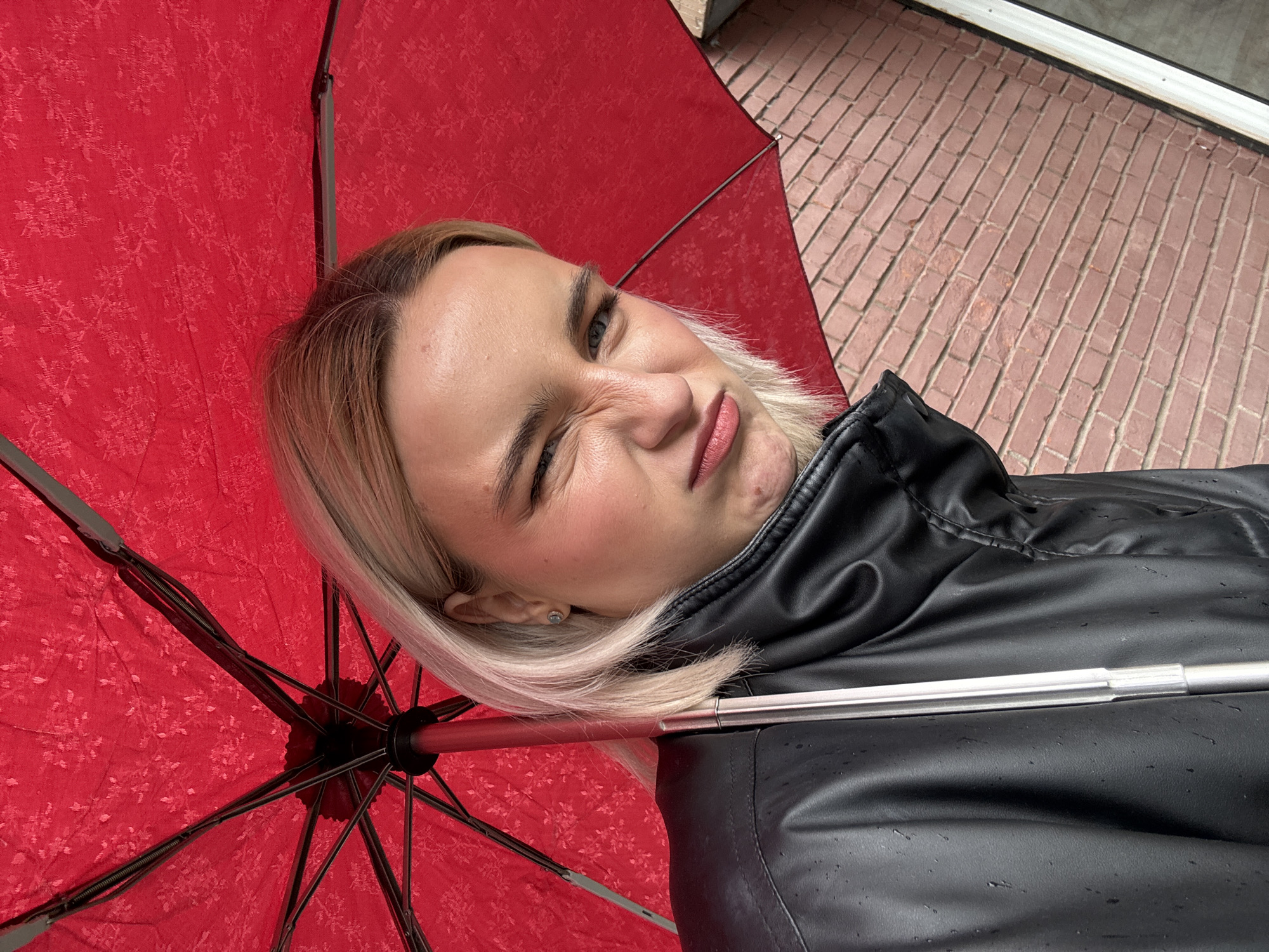 The image shows a person holding a red umbrella in Saint Petersburg, Russia. The person, who appears to be female, is wearing a black leather jacket and has blonde hair. Her face is angled toward the camera, and she is winking with one eye. The background features a brick-paved area, possibly a sidewalk or pedestrian zone, and the edge of a building with a window.
