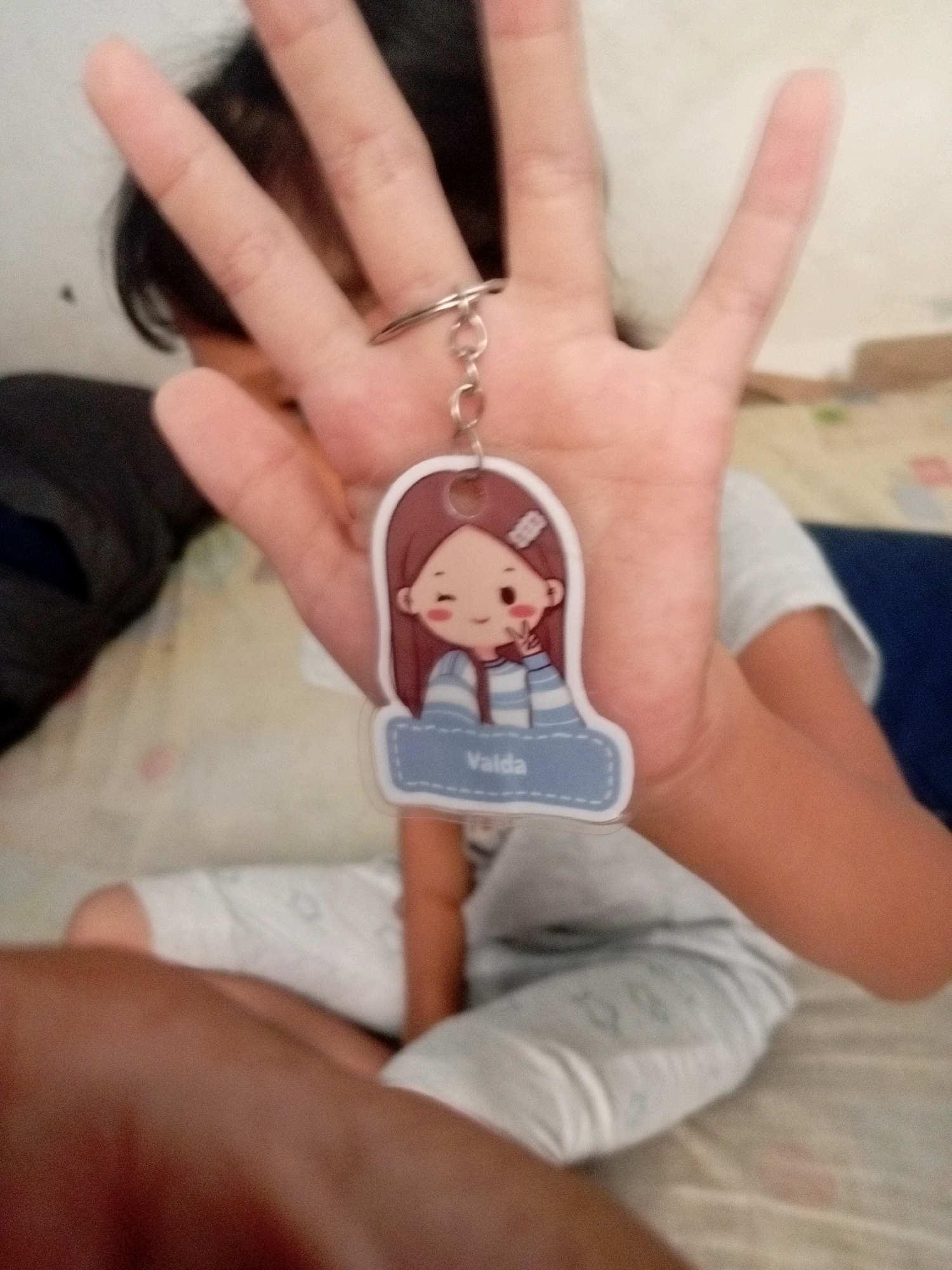 A close-up, slightly blurry image shows a person's hand holding a keychain. The keychain features a cartoon character of a girl with brown hair, wearing a blue and white striped shirt, and making a peace sign with her fingers. The character's name, "Valda," is written on a blue banner below her. The hand holding the keychain appears to be that of a child. The background is out of focus, but suggests an indoor setting, possibly a bedroom, with bedding visible. There is no information in the image that indicates the location in Batam, Indonesia.