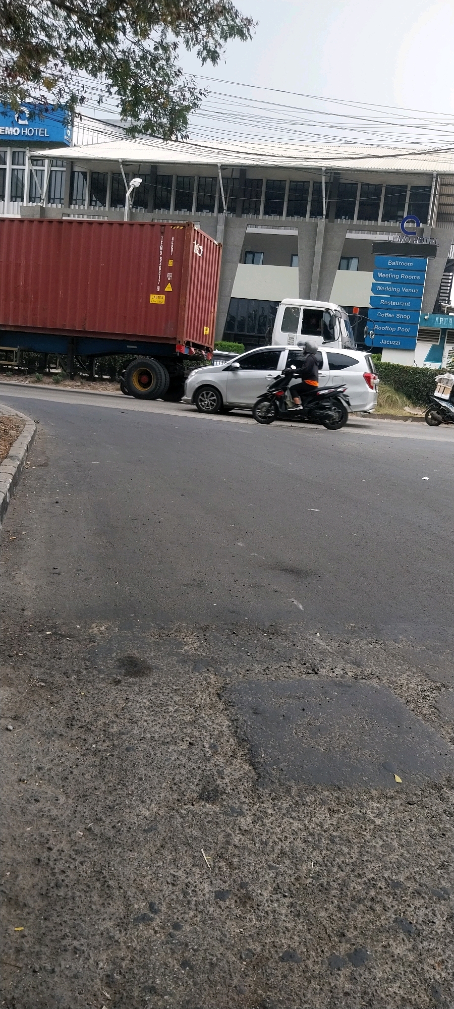 The image depicts a traffic incident on a road in Kelapa Dua, Indonesia. In the foreground, a portion of a damaged asphalt road is visible, with several potholes and patches of repair. To the left, a concrete curb runs along the edge of the road. In the background, a large red shipping container is being transported on a truck, its rear wheels visible. The container appears to be partially detached from the truck, suggesting a potential accident. To the right of the container truck, a silver multi-purpose vehicle (MPV) is positioned, with its front end close to the container. A black scooter with a rider wearing a helmet and an orange shirt is in motion, positioned between the MPV and the shipping container, suggesting it may be attempting to maneuver around the obstruction. Further in the background, a white truck cab is partially visible behind the MPV, its front window showing a person. The Lemo Hotel is prominently displayed on a sign in the background, along with smaller signs indicating various amenities such as "Ballroom," "Meeting Rooms," "Wedding Venue," "Restaurant," "Coffee Shop," "Rooftop Pool," and "Jacuzzi." The sky is clear and bright, indicating a daytime scene. Trees with green foliage are visible on the left side of the image, and power lines are strung across the upper portion of the frame. The overall scene suggests a disruption of traffic flow due to the apparent difficulty or failure in transporting the shipping container.
