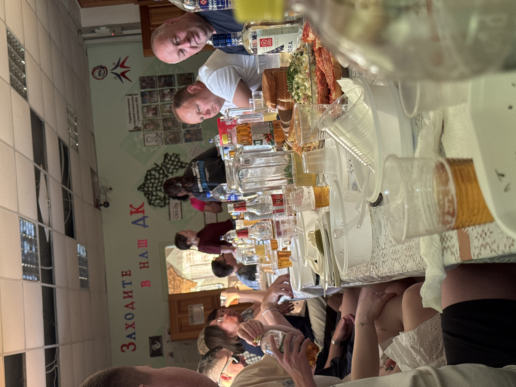A medium shot depicts a group of individuals gathered around a table laden with food and beverages, appearing to be at a celebratory event. The location is indicated by the inscription "Заходите в наш АК" (Come to our AK), suggesting the event is taking place within a setting related to "AK," potentially a cultural or local community center. The interior features a wall adorned with various images and text in Russian, complemented by a wall mural that adds to the festive ambiance.
The participants range from middle-aged to elderly, creating a heterogeneous demographic that spans different age groups. They are engrossed in eating, drinking, and conversing, indicating the social aspect of the gathering. The table serves as a focal point, showcasing a wide array of Russian food and drinks, including salads, breads, and multiple bottles of vodka and other spirits, highlighting the cultural emphasis on hospitality and communal dining.
The subjects and objects, in conjunction with the people's actions, clearly suggest that the picture was taken at an event commemorating a significant event in Кемуль, Russia.