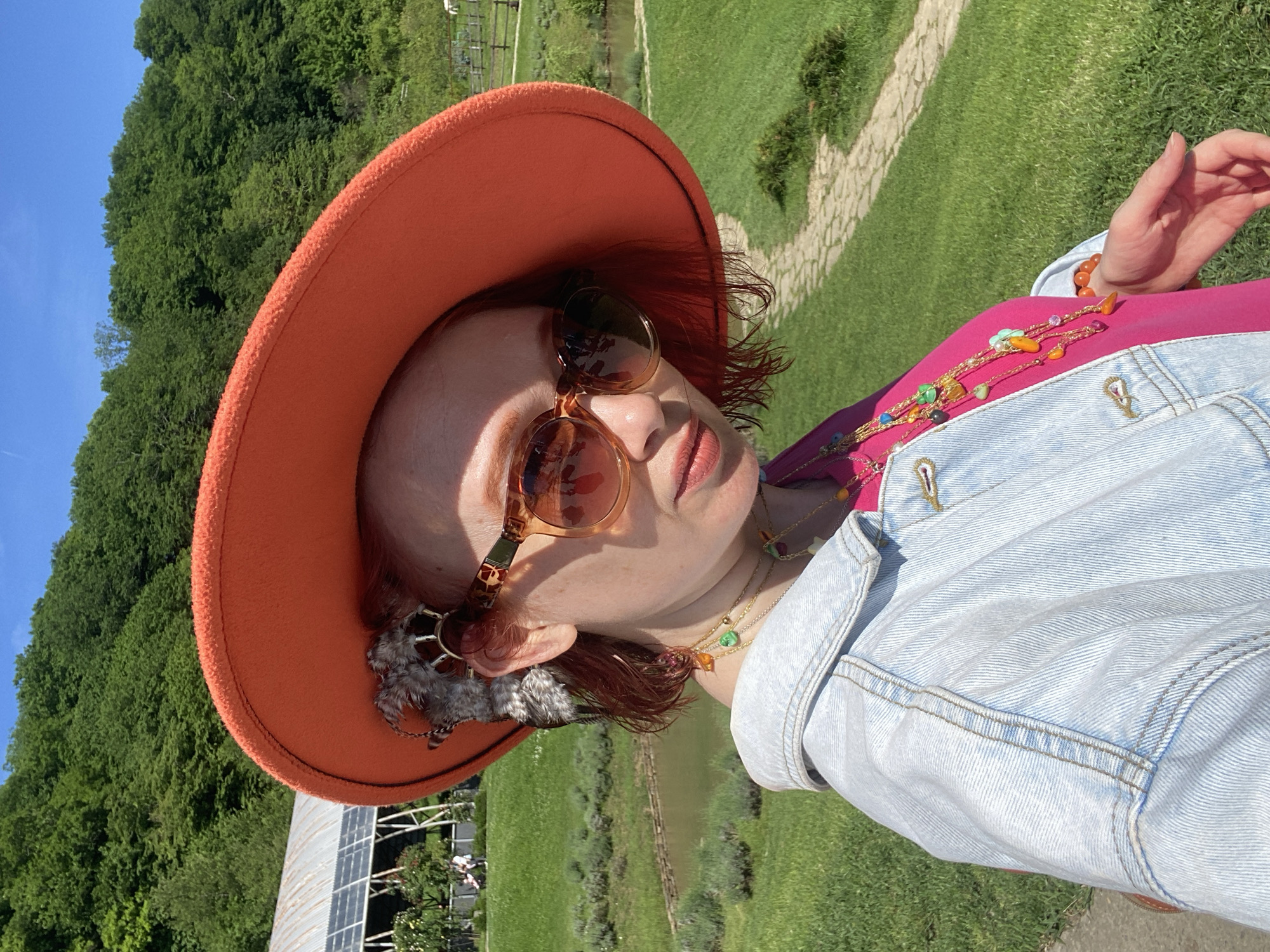 A woman wearing an orange hat and sunglasses stands outdoors. She also wears a denim jacket over a pink shirt, accessorized with several necklaces and a feather earring. The background consists of green grass, trees, a body of water, and a stone path. Solar panels and a building are partially visible in the lower left corner. The location is not identified as being in Прогресс, Russia.