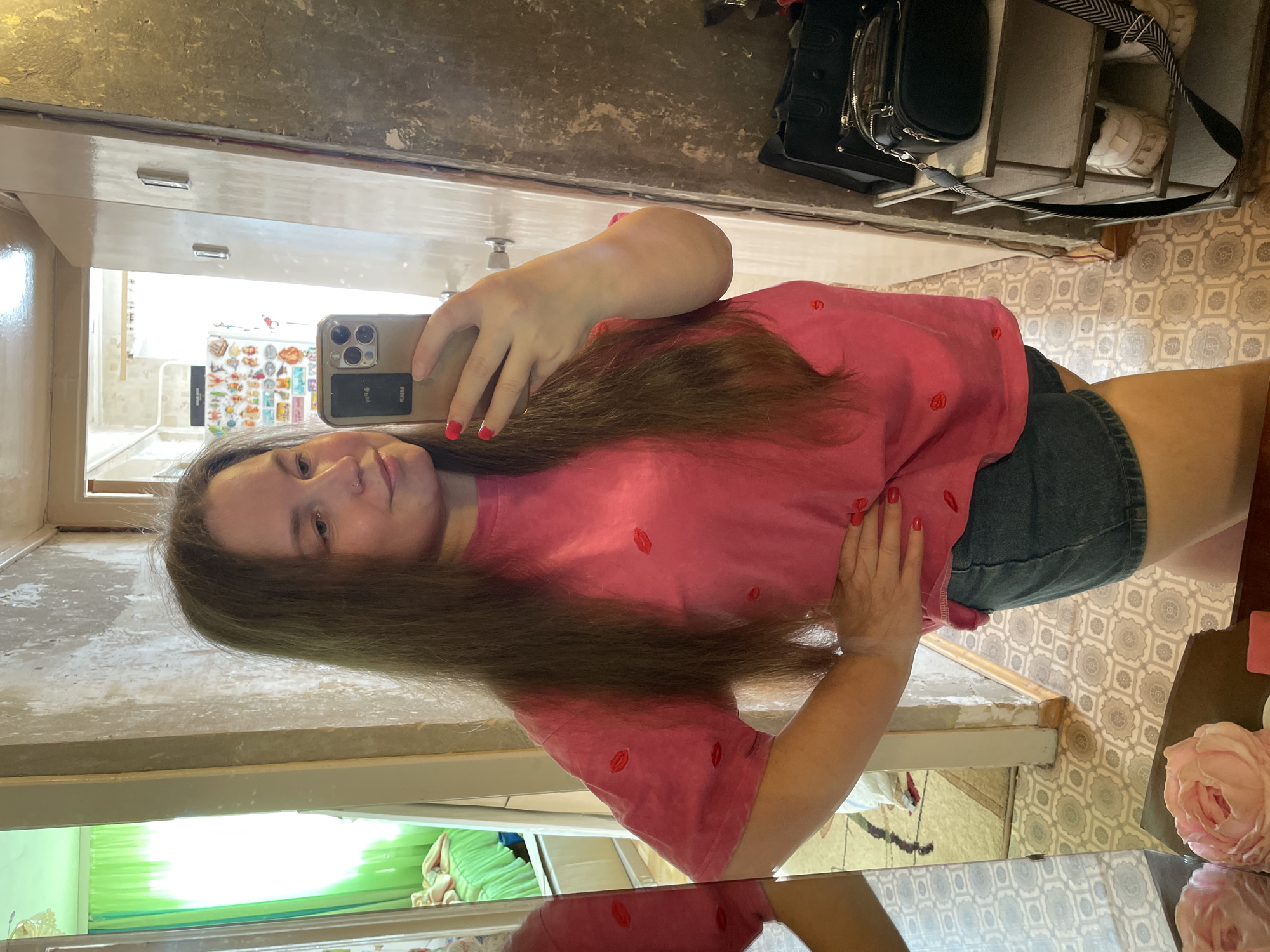 A woman is taking a selfie in a mirror. She is wearing a pink t-shirt with red lip prints and denim shorts. The room has light-colored walls and a patterned floor. Reflected in the mirror is a glimpse of another room with a green curtain. In the background, there is a shelf with bags and shoes. The woman has long brown hair and is holding a phone with multiple lenses. The location appears to be inside a building, but the specific city (Izhevsk, Russia) is not discernible from the image.