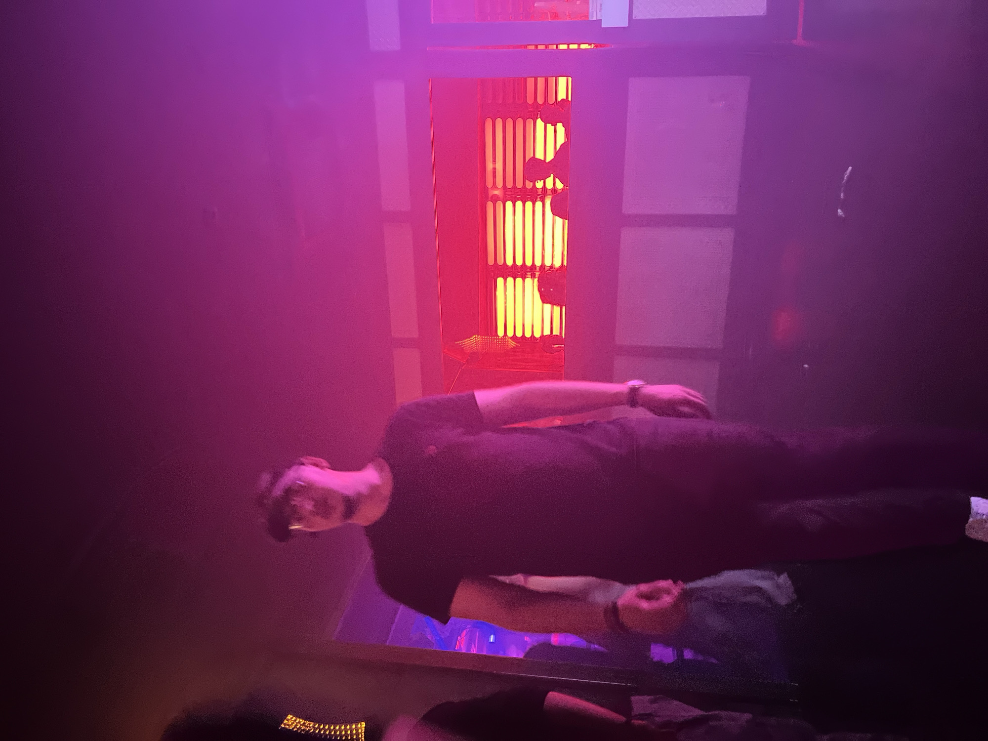 A man is lying on a surface in a room bathed in pink and red lighting. The man is wearing dark clothing. An illuminated object with a grid-like pattern and red and yellow light stands vertically in the background. The location is Strasbourg, France.