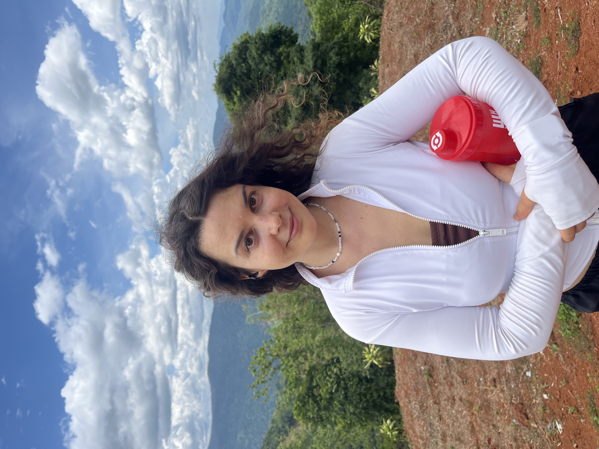 A woman with curly brown hair stands outdoors holding a red water bottle. She is wearing a white zip-up jacket over a brown top and black shorts. She has on a beaded necklace. Behind her is a mountainous landscape with green trees and a blue sky with white clouds. The ground in front of her is reddish-brown dirt with sparse vegetation.