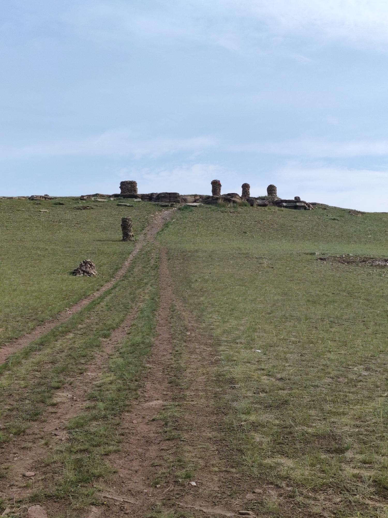 A dirt path winds up a grassy hill in Askiz, Russia, leading to a collection of ancient stone pillars. The sky is a pale blue with scattered clouds. On the left side of the path, a small pile of stones rests on the grass. Further up the hill, near the path, stands a single, tall stone pillar. At the summit of the hill, a cluster of several more stone pillars are arranged amongst rocks and sparse vegetation. The overall scene depicts a historical or archaeological site in a natural, open environment.