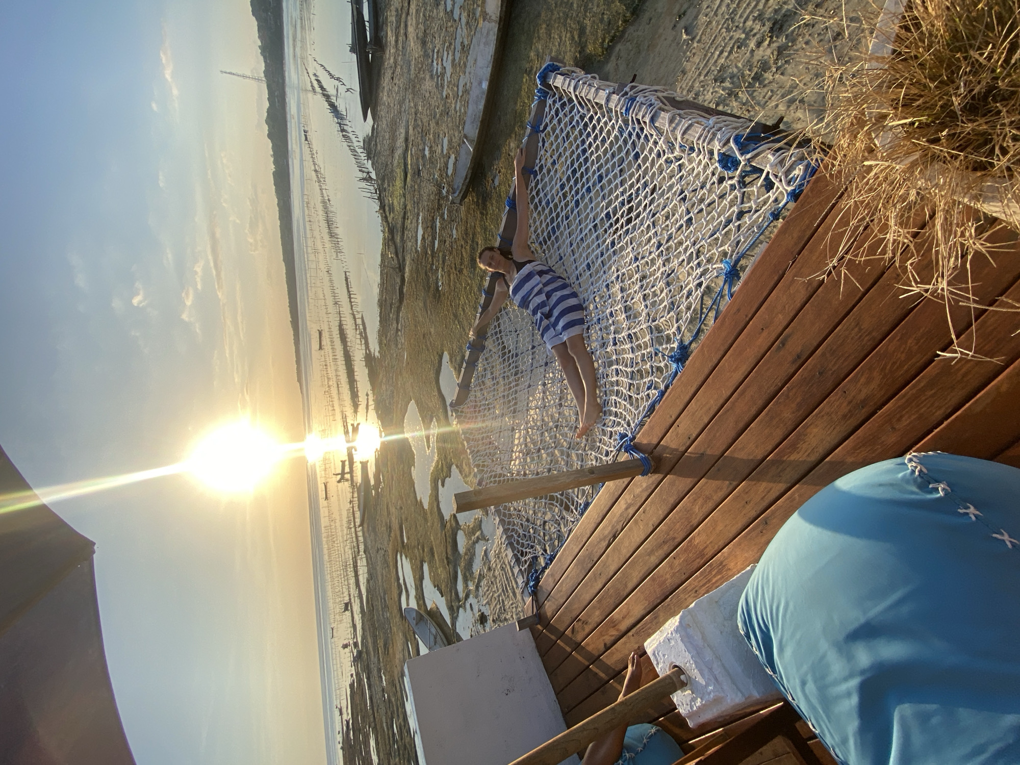 This is a photograph taken in Nusa Penida, Indonesia, depicting a woman enjoying a hammock at sunset. The woman, wearing a blue and white striped dress, is positioned in the hammock, stretching her arms and legs. She appears relaxed and happy.
The setting is a beach with shallow water, sand, and numerous wooden posts or pilings protruding from the water, suggesting a traditional fishing or aquaculture setup. The sun is low in the sky, casting a warm, golden light and creating a lens flare effect. This indicates that the photo was taken during the golden hour, likely just before sunset. The sky is clear with a few wispy clouds.
The hammock is made of white netting with blue accents and is suspended over a wooden deck. In the foreground, parts of the wooden deck and a large blue beanbag chair are visible. The overall atmosphere is serene and idyllic, conveying a sense of vacation and relaxation. There is no visible text in the image.