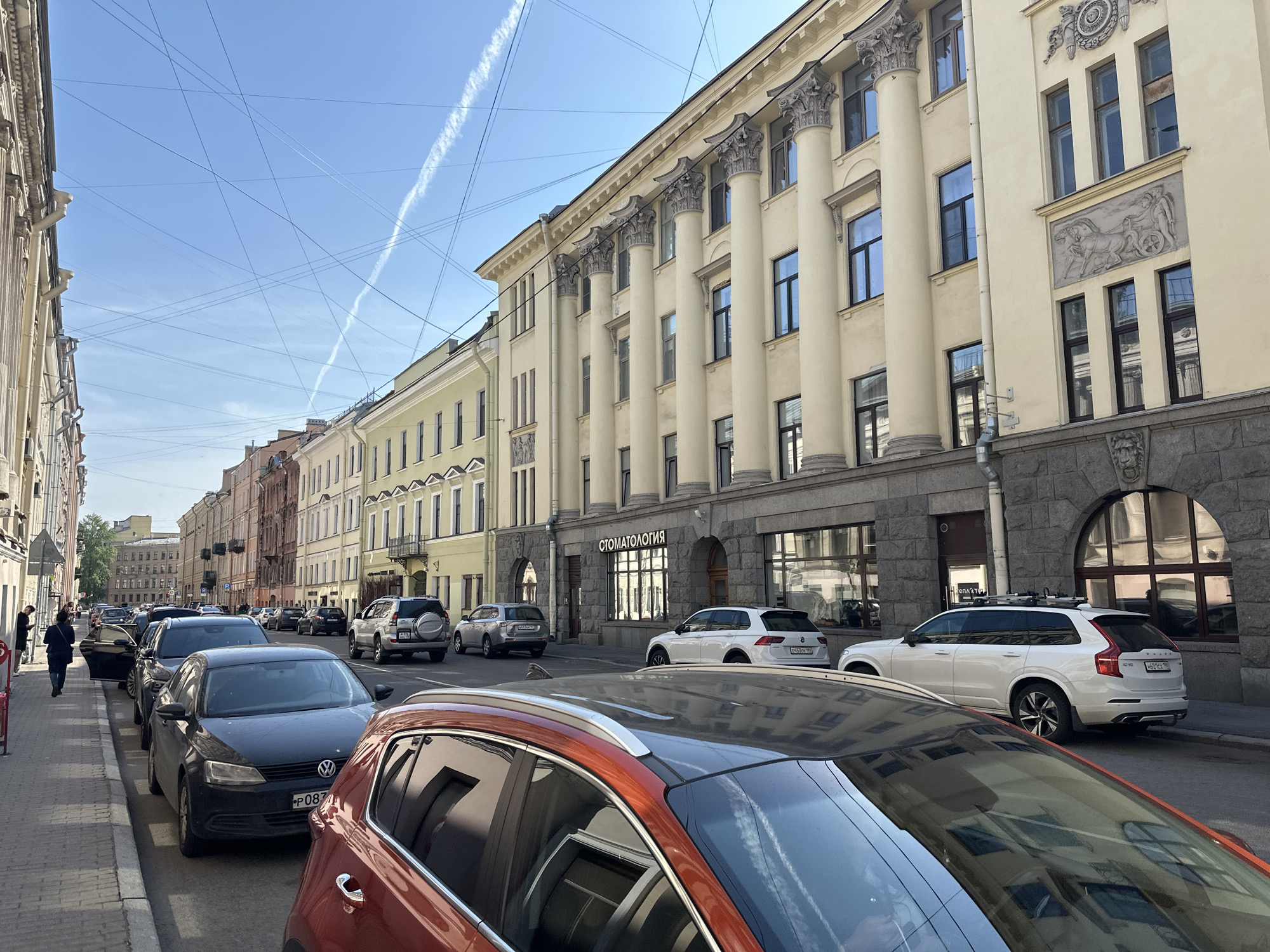 The image shows a street in Saint Petersburg, Russia, lined with parked cars on the right side. Pedestrians walk on the sidewalk to the left. The buildings on the right are characterized by neoclassical architecture, with columns and ornate facades. A dental clinic ("СТОМАТОЛОГИЯ") is located on the ground floor of one of the buildings. Overhead power lines and contrails crisscross the sky. The image depicts a typical urban scene in Saint Petersburg.