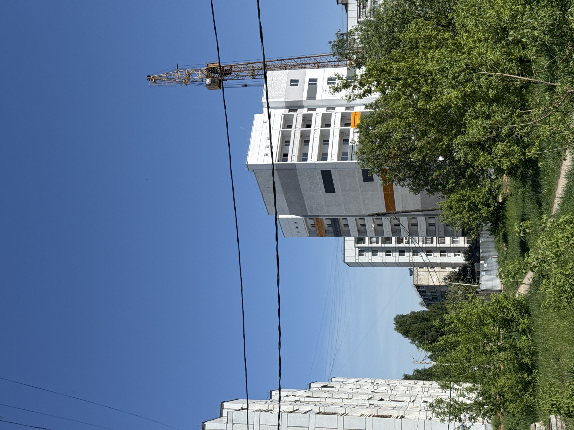 The image shows a partly cloudy blue sky over a construction site in what is likely Chaikovsky, Russia. A tall crane is visible near the top of a white, modern building under construction. Utility lines run across the bottom left of the frame. Green trees and some grass can be seen to the right of the building, and another structure, possibly an apartment building, is situated at the bottom of the image. The photograph is rotated 90 degrees clockwise.
