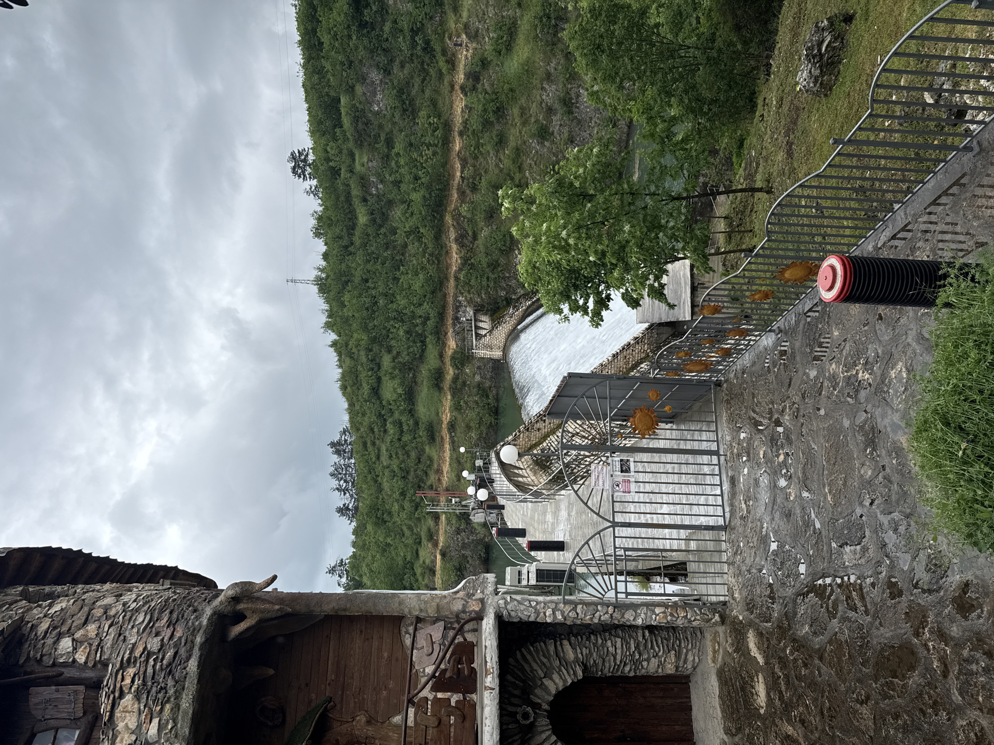 The image shows a landscape scene in Lukovit, Bulgaria, featuring a stone building with a wooden door in the foreground. A man-made waterfall flows behind a metal gate. The water flows out of a river between densely forested slopes. A pedestrian bridge with lights spans the river, connecting to another gate on the far side. The sky is overcast. The scene is viewed from an elevated position looking down towards the waterfall. The architecture incorporates natural stone elements.