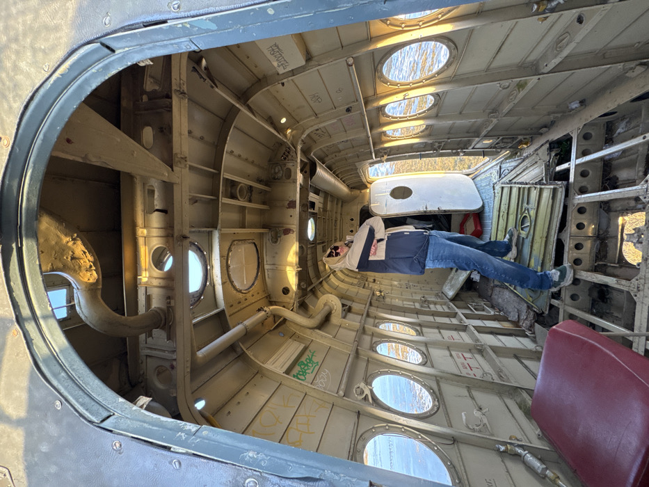 The image depicts the dilapidated interior of an aircraft fuselage located in Падлабенскі сельскі Савет, Belarus. The light-colored walls show signs of wear, dirt, and graffiti in green and black markings. Exposed structural ribs, piping, and wiring are visible along the ceiling. The cabin floor is largely absent, revealing the underlying metallic framework and debris. A male individual stands in the mid-cabin, facing slightly left, with his head tilted upwards and arms crossed. He is wearing a two-tone hooded jacket (beige top, dark blue bottom), blue jeans, sunglasses, and green/white sneakers. Multiple circular windows line both sides of the fuselage, showcasing a sunlit body of water outside. An open door on the starboard side provides a view of an exterior grassy bank bordering the water. A red upholstered seat cushion is present in the lower left foreground.