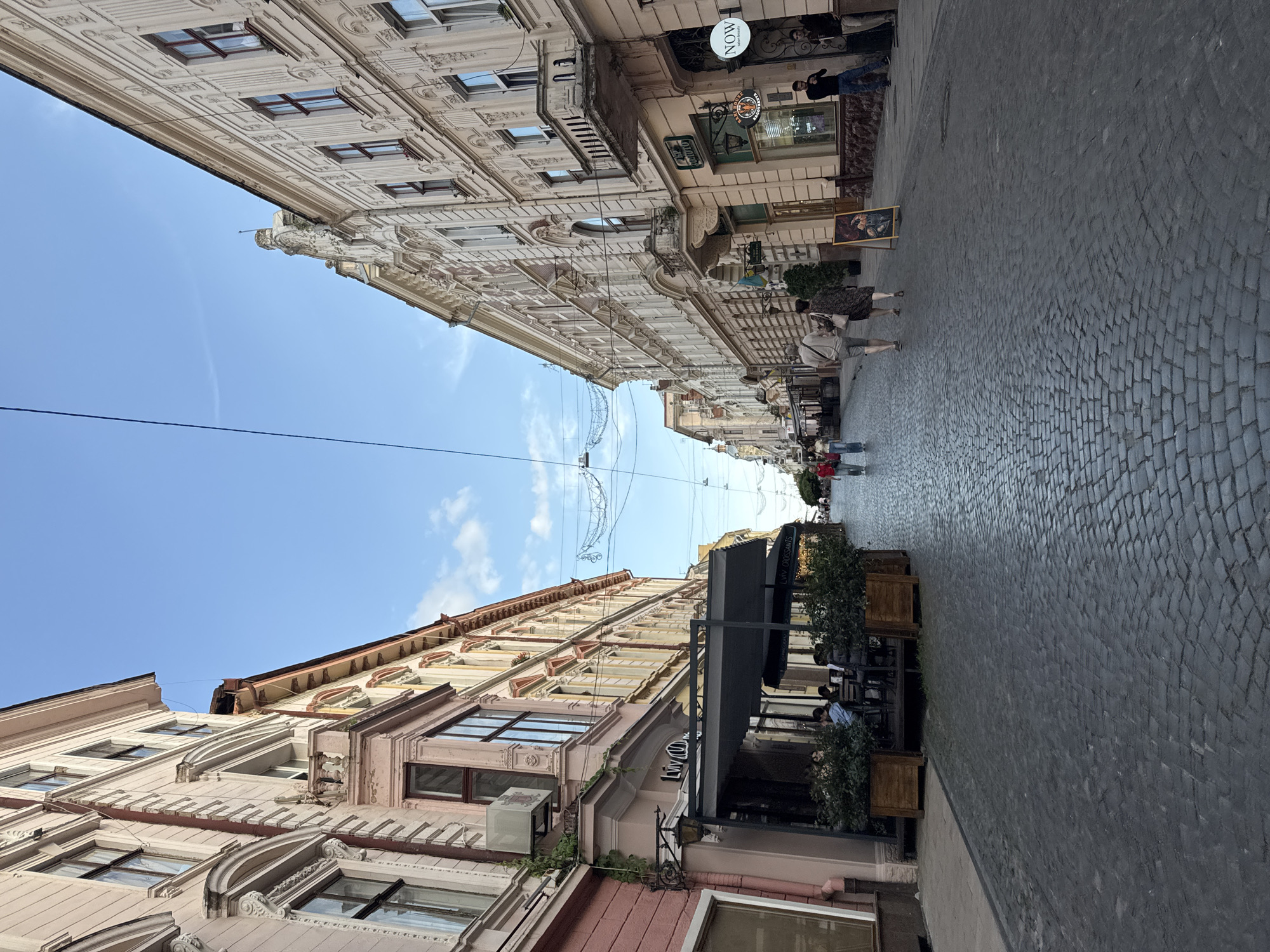 The image depicts a street scene in Chernivtsi, Ukraine, likely on a clear day with a few wispy clouds. The street is paved with cobblestones, and on either side, there are multi-story buildings with ornate architectural details. These buildings appear to be a mix of residential and commercial spaces, with awnings and signs indicating shops and cafes. Several people are visible walking along the street, some sitting at outdoor cafe tables, and others standing near storefronts. Overhead, power lines and decorative string lights are strung across the street. The overall impression is of a bustling urban environment with historical architecture.