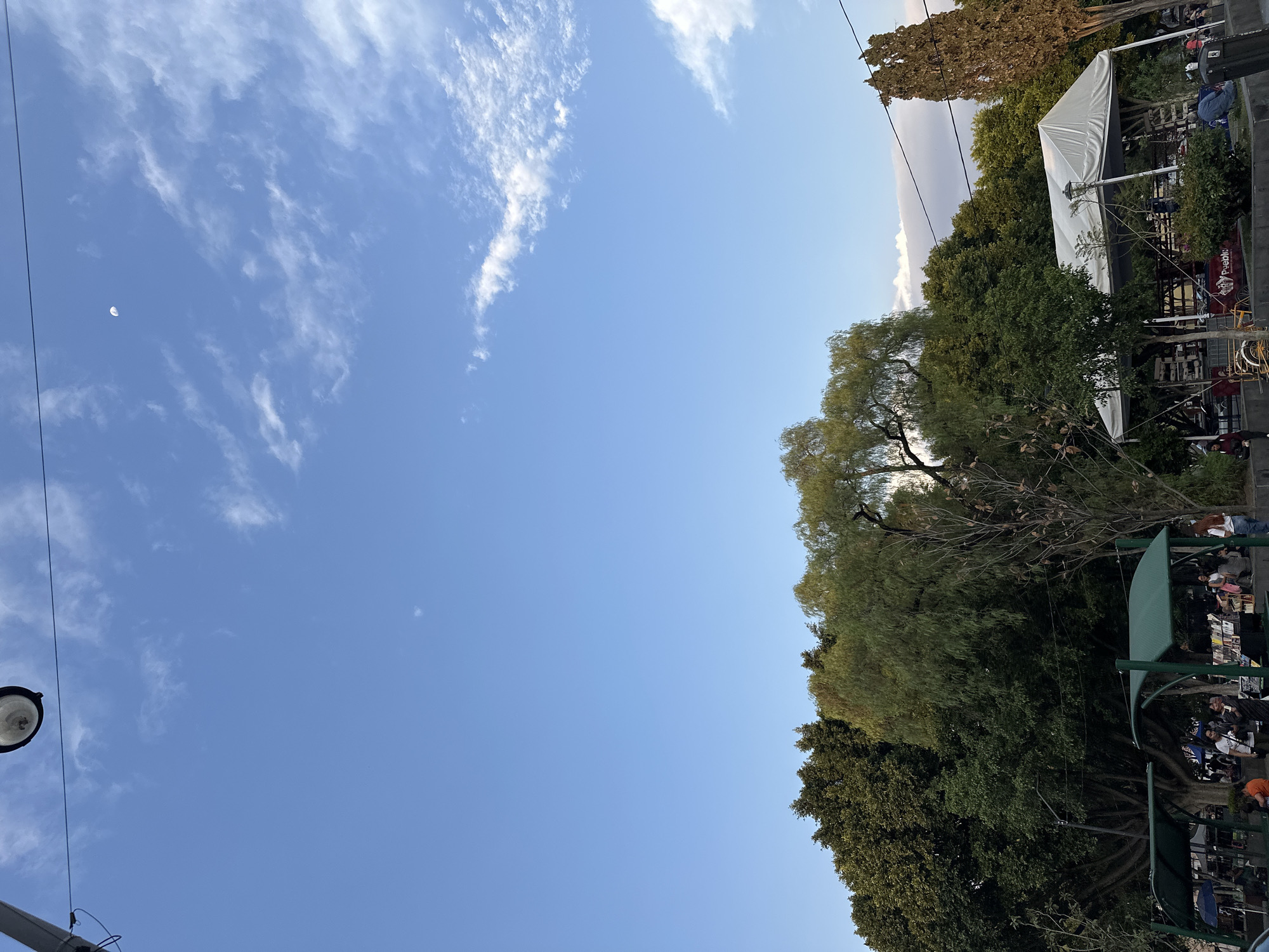 The image captures a scene in an urban area of Unknown, Unknown, showing a partly cloudy sky with a crescent moon visible. The lower right portion of the image is dominated by lush green trees. There's a glimpse of what appears to be an outdoor seating area, possibly a restaurant or café, with white canopies. There are people visible sitting under the green awnings. On the lower left is a streetlight with wires.