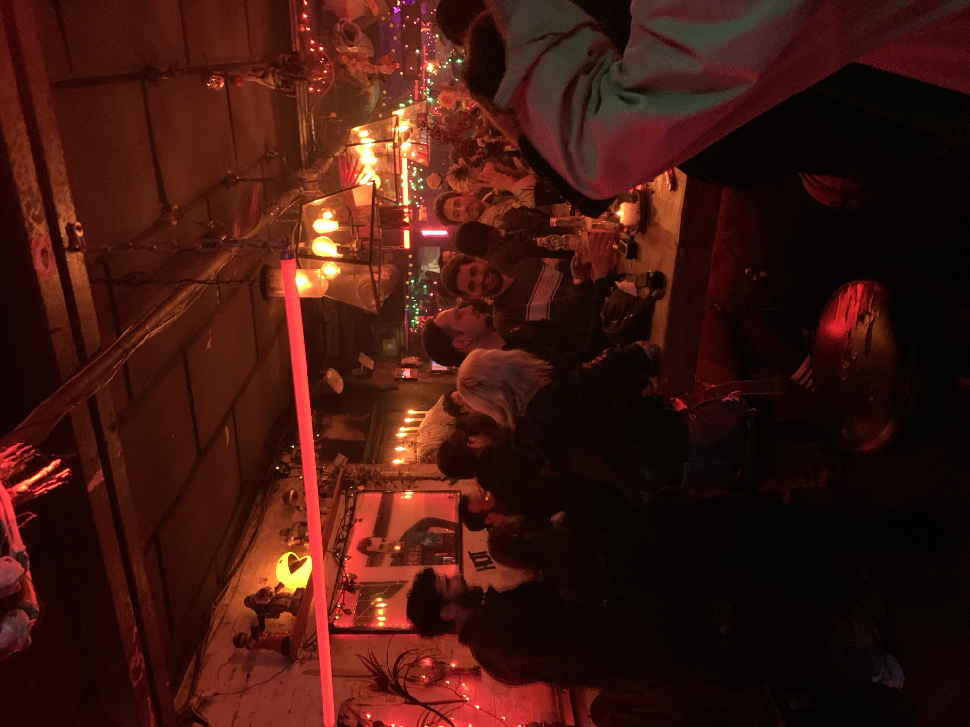 The image depicts an indoor scene at a bar or restaurant in Tel Aviv, Israel. Several people are seated at a table, possibly engaged in conversation or having a meal, amid a dimly lit setting. Overhead, there are exposed pipes and beams, decorated with string lights and other festive decorations. On the wall, artwork or photographs are visible, with various decorative elements surrounding them. The lighting is predominantly red and orange, with candles and neon lights contributing to the ambiance. The location is a crowded establishment, suggesting a lively social gathering.