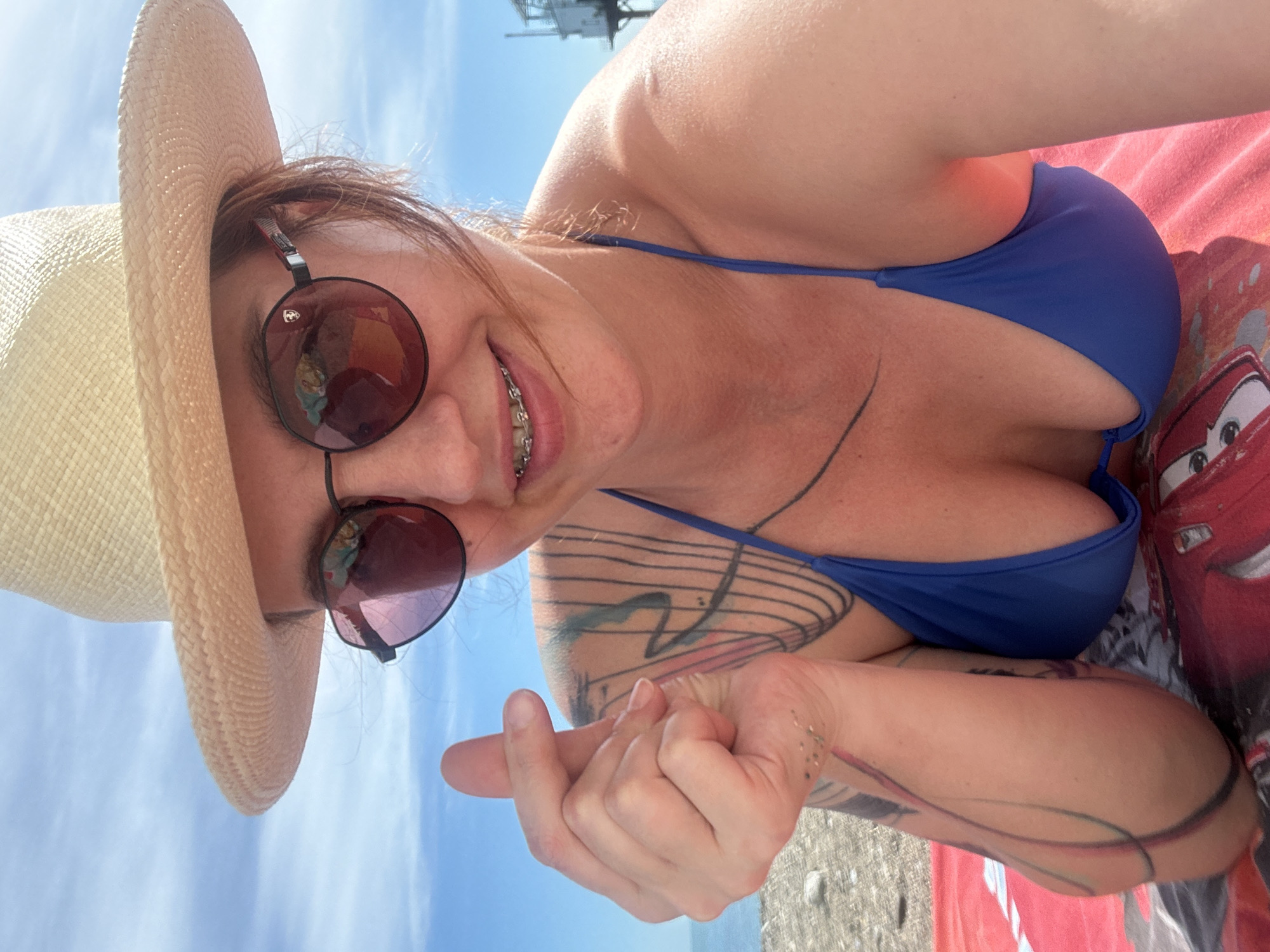 A woman is captured in a tilted, close-up selfie, likely at a beach in Sochi, Russia. She is wearing a light-colored straw hat and round, dark sunglasses. Her dark hair peeks out from under the hat, and she is smiling broadly, revealing dental braces. She wears a blue bikini top, which shows off several tattoos on her arm and shoulder. One tattoo appears to be a series of lines or musical notes.
With her left hand, she makes a finger heart gesture, pointing it towards the camera. She is reclining on a red blanket or towel featuring a recognizable animated character, Lightning McQueen from Disney Pixar's "Cars." The background shows a clear, bright blue sky, suggesting sunny weather. In the distance, a dark structure, possibly a pier or part of a coastal building, is visible against the sky. The ground beneath her appears to be a mix of sand and small pebbles, characteristic of a beach. The overall mood is relaxed and joyful.