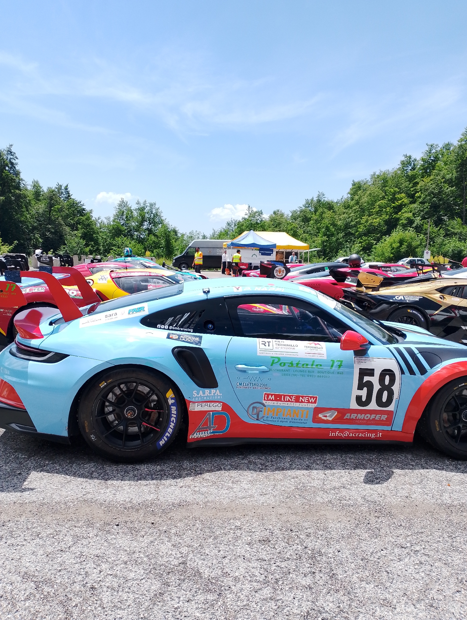 The image shows a row of colorful race cars parked on asphalt, with a backdrop of lush green trees and a bright blue sky with scattered clouds. In the foreground, a light blue Porsche 911 GT3 Cup car with red and black racing stripes is prominently displayed. The car has the number "58" in large black digits on its side, along with various sponsor logos, including "SARPA COSTRUZIONI," "PEREGO," "LM - LINE NEW ARREDAMENTI RUSTICI," "IMPIANTI," "ARMOFER," and "info@acracing.it."
Behind this car, several other race cars are visible, including a yellow car, a pink car, and a red car with a large spoiler. Tents with yellow and blue fabric are set up in the background, suggesting an event or race. A few people are present, including one person in a yellow vest standing near a black van and others standing around the tents. The overall scene suggests a motorsport event taking place in an outdoor, possibly rural or semi-rural setting, given the surrounding trees. While the image doesn't explicitly state the location in Rieti, Italy, one of the visible sponsor logos on the car reads "RIETI TERMINILLO O2." This suggests the event is likely taking place in or near Rieti, possibly at the Terminillo circuit or a related venue.