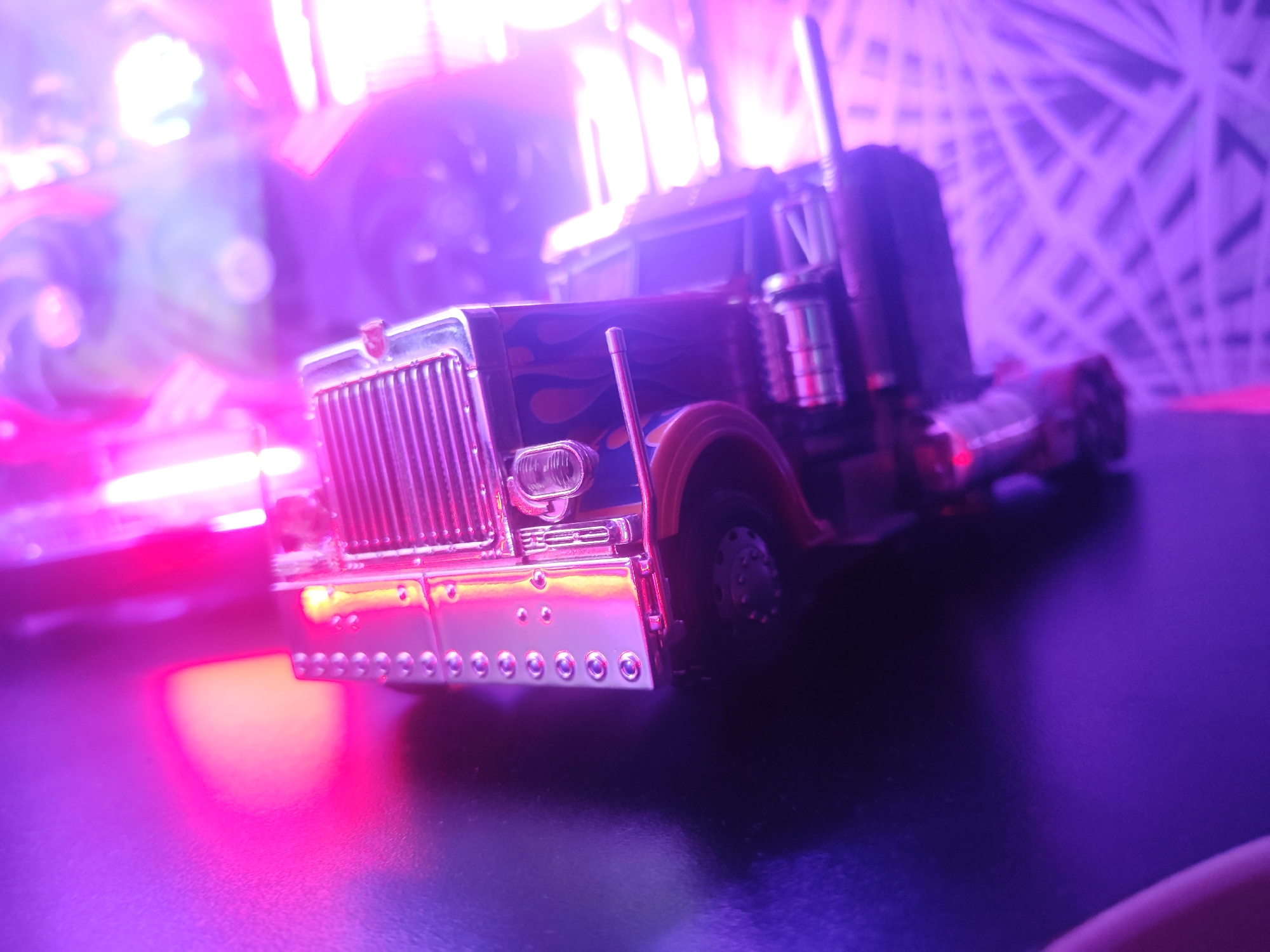 An extreme close-up shot focuses on the front grille and hood of a toy semi-truck. The truck is dark blue with stylized orange and yellow flames painted on the side of the hood. The grille and bumper are made of shiny chrome, reflecting the purple and pink lights that illuminate the scene. Several small circular silver studs are visible along the bottom edge of the bumper. A portion of the driver's side front tire is visible to the right of the truck. The background is heavily blurred and bathed in the same purple and pink light, with some geometric shapes suggested in the upper right portion of the image. There is no indication of the city of Borova, Ukraine.