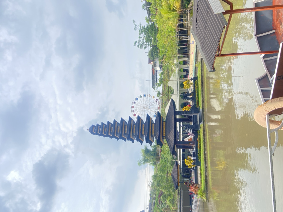 The photo depicts a tall, tiered, dark-colored Asian temple structure, possibly a pagoda. A Ferris wheel is visible in the background. The temple is surrounded by greenery and situated near a body of water that seems to be a man-made canal or pond. A covered structure appears on the right side of the water, possibly a boat or dock area. The sky above is overcast, with clouds filling a large portion of the frame.