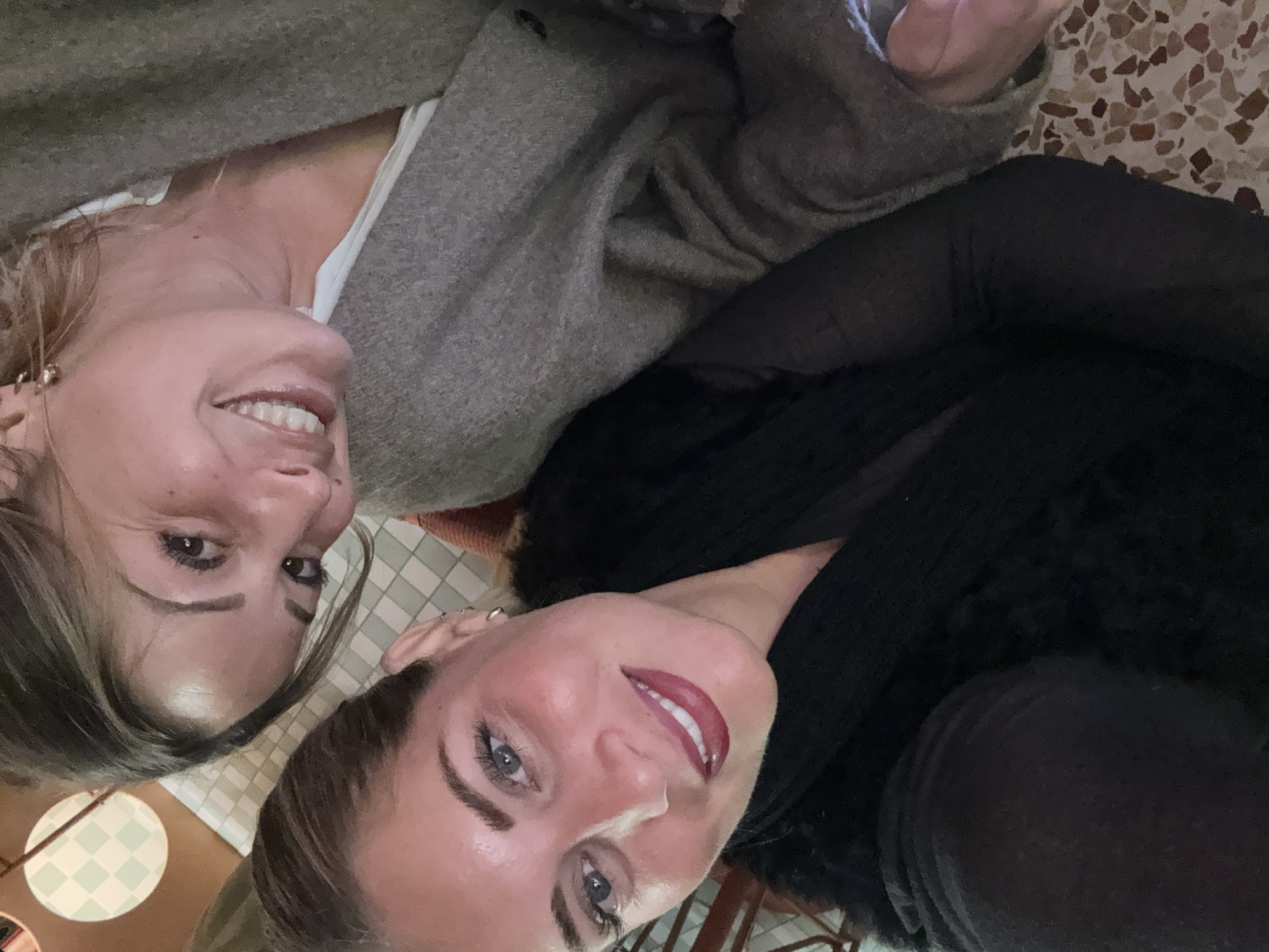The image shows two smiling women in an indoor setting. One woman is wearing a light-colored shirt and a gray sweater. The other woman is wearing a black outfit. The background includes a table with a checkered pattern and a floor with a patterned surface. There is no clear indication of Croix, France.