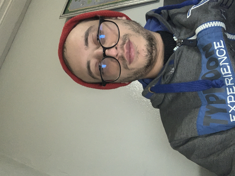 A person wearing a red beanie and glasses poses for a selfie indoors. The subject has a beard and is wearing a grey and blue hooded sweatshirt with the words "TYPHOON EXPERIENCE" printed on the front. The background includes a beige wall and a framed picture, suggesting a room interior. The person is positioned at an angle relative to the camera.