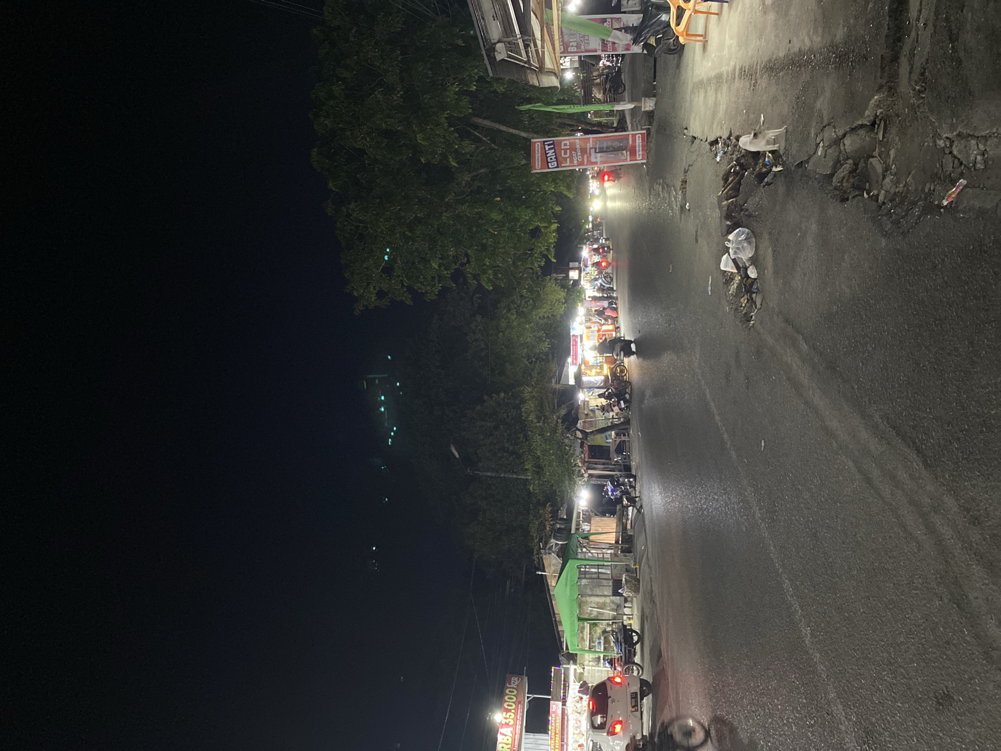 The image depicts a nighttime scene on a street in Banda Aceh, Indonesia, likely a commercial area. A white car is parked on the left side of the road, with a street vendor stall behind it. Further down the road, several other stalls and shops are visible, illuminated by artificial lights, with motorcycles and people present. Trees line the right side of the street, and a large sign for "GANTI LCD" (meaning "Change LCD" in Indonesian) is prominently displayed. The road surface shows signs of damage and debris, with trash scattered near a large pothole. A dog stands near the damaged road. The overall atmosphere suggests a lively but somewhat unkempt street at night.
