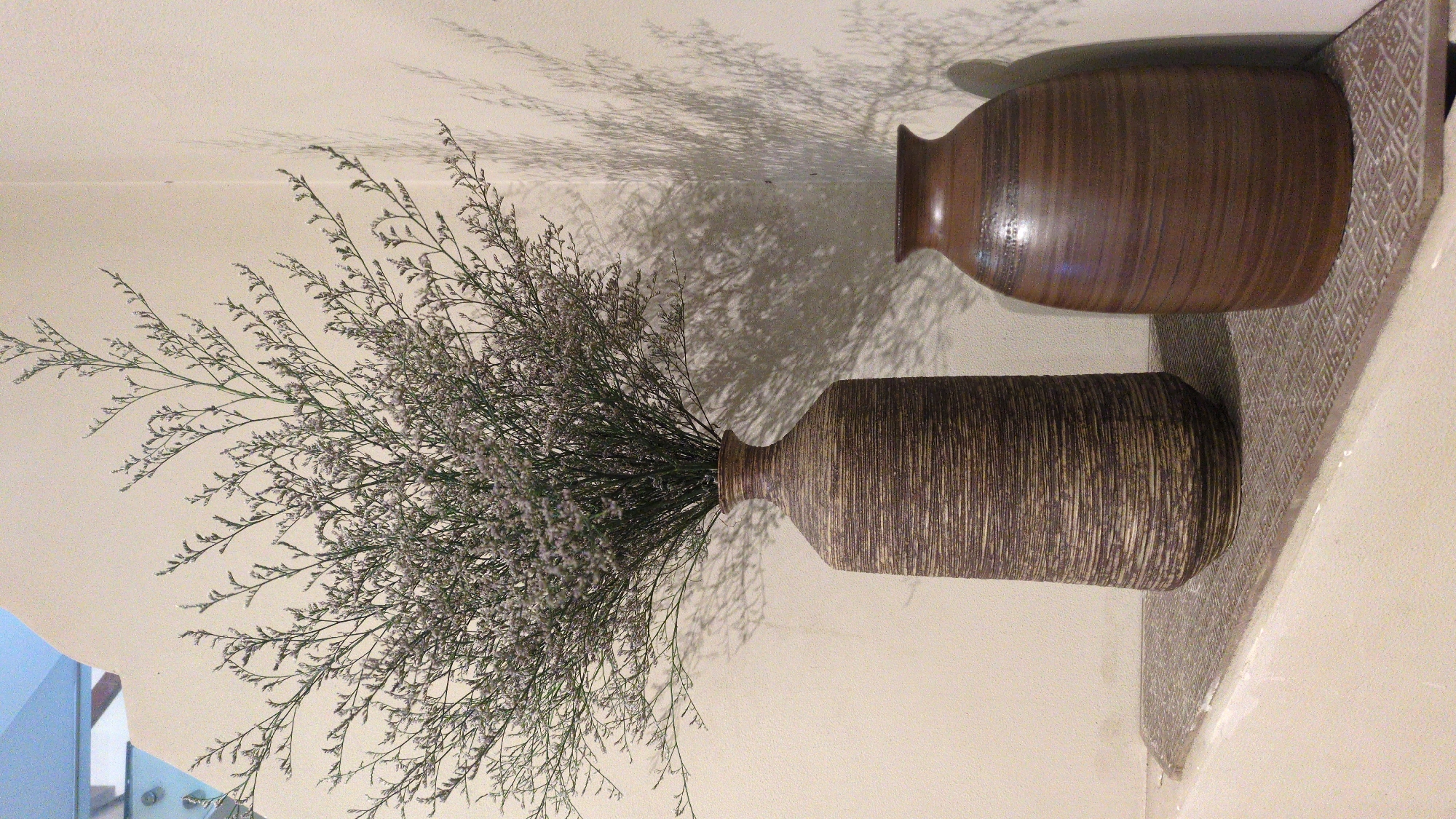 Two vases sit on a shelf against a light beige wall. One vase holds a spray of dried, wispy flowers and casts a shadow on the wall. The vases are brown with horizontal ridges. The shelf is decorated with a geometric pattern. The photo was likely taken in Tuy Hòa, Vietnam.