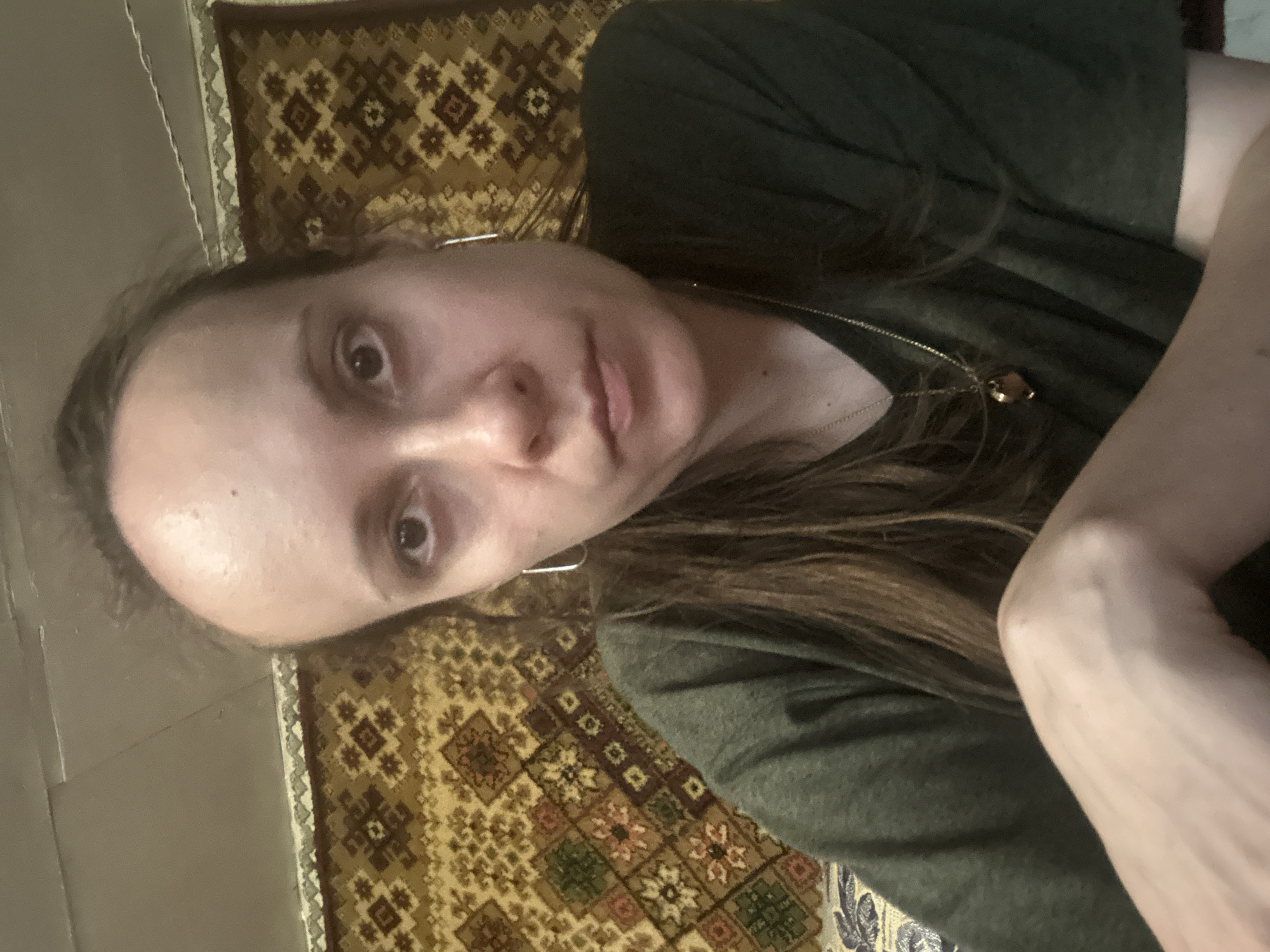 The media file is a selfie of a person lying down on a patterned rug. The person has long brown hair and is wearing a dark green shirt with a necklace. Only the upper part of the person's body is visible, including their face, arm, and shoulder. The person's expression is neutral. The lighting suggests it is daytime, but there are no external cues about the specific location or weather. The rug has a detailed pattern with geometric shapes and floral motifs in shades of brown, yellow, and green. The background includes a neutral-colored wall. There is no visible text in the image.