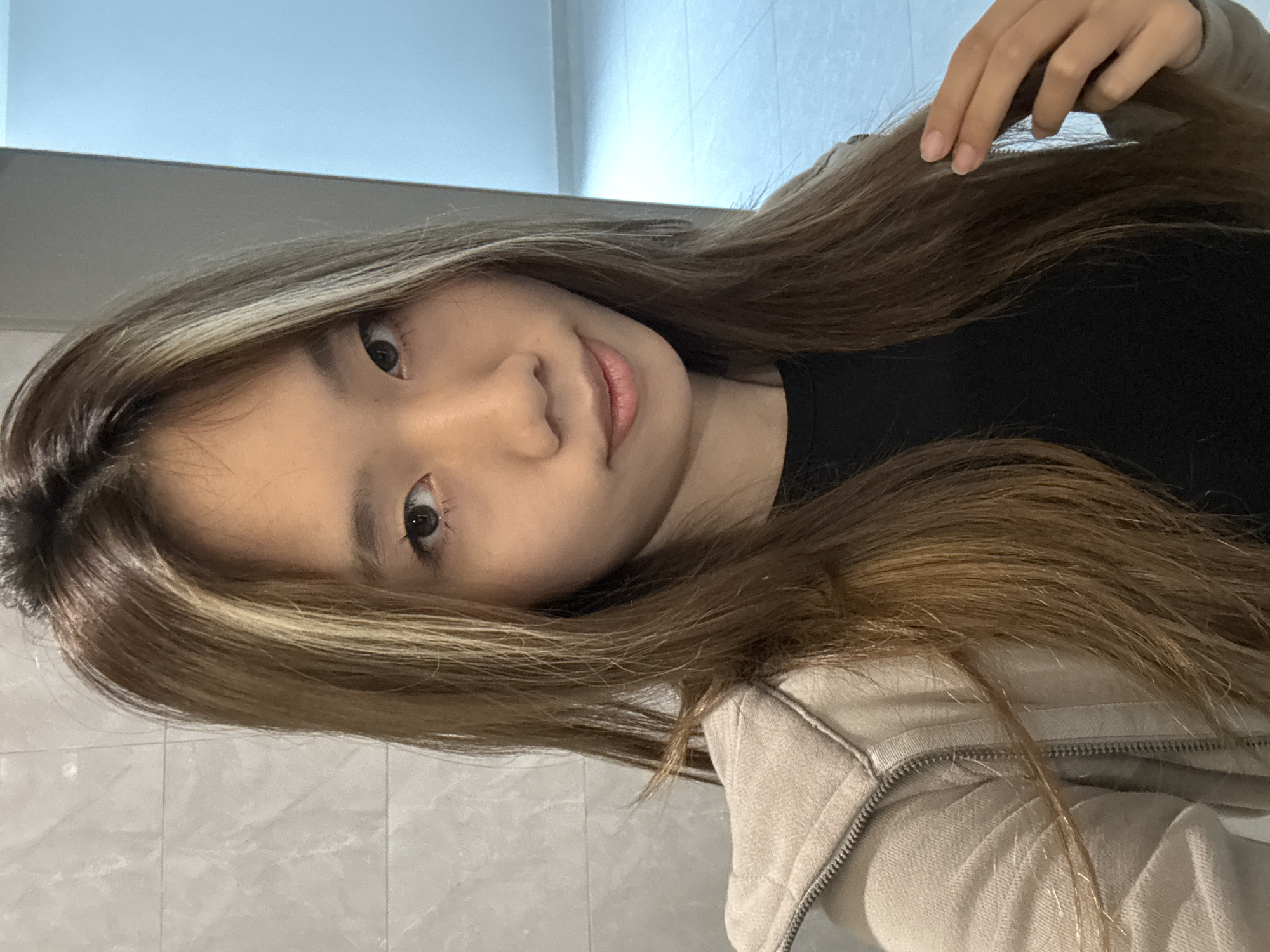 A young Asian woman is captured in a close-up, chest-up shot, looking directly at the camera with a soft, gentle smile. Her head is tilted slightly. She has long, straight hair, medium brown with prominent lighter blonde or caramel highlights framing her face and running through the length of her hair. She is wearing a black top under a light beige or off-white zip-up hoodie, which has a visible zipper and ribbed trim. Her right hand is visible near her head, with natural, unpainted fingernails, seemingly touching or adjusting her hair.
The setting appears to be indoors, likely in Nan'an District, China. The background features a light-colored, possibly white or light grey, tiled wall below, and a plain, lighter-toned wall or ceiling area above. The lighting is soft and even, suggesting indoor artificial illumination or diffused natural light. No text is visible in the image.