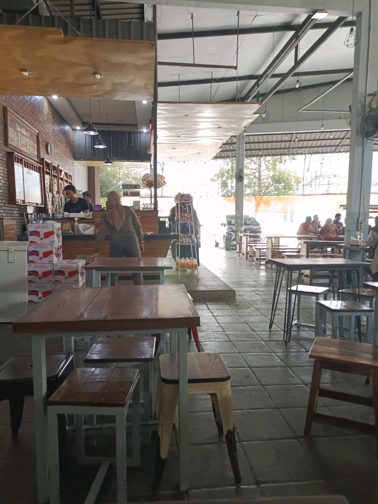 The image depicts an indoor/outdoor cafe or food stall setting in Made, Indonesia. The foreground features several wooden tables and stools, with a focus on one table in the lower center with a light blue metal frame and a wooden top. To the left, stacks of boxes with red, white, and blue graphics are visible near a counter area. In the background, several people are seated at tables, engaged in conversation or eating. A person wearing a headscarf and a khaki-colored jacket is standing behind a counter, interacting with items on display. Another person, a man with short dark hair, is visible behind the counter, also appearing to be working. The environment appears to be covered by a corrugated metal roof supported by metal beams, with open sides allowing for natural light and views of trees and vegetation outside. The flooring is tiled. The overall impression is of a casual, communal dining space.