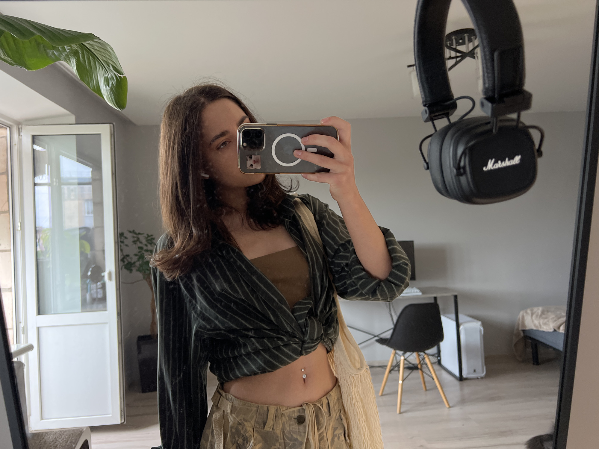 A person is taking a mirror selfie in a room, reflected in a large mirror. The person is wearing a green striped, knotted shirt, a brown bandeau top, and camouflage pants. They have a belly button piercing. On the left side of the mirror, a large green leaf from a plant is visible. Above the person's reflection, a pair of black Marshall headphones are hanging. In the reflection of the room, there is a desk with a computer monitor, a white computer tower, and a black chair with wooden legs. A bed with a beige blanket is visible in the background. A white door with glass panes is also in the reflection. The flooring appears to be light-colored wood laminate. The overall environment suggests an indoor personal space, possibly a bedroom or living area, within a residential setting. Given the metadata of the image, the location is identified as Ternopil, Ukraine.