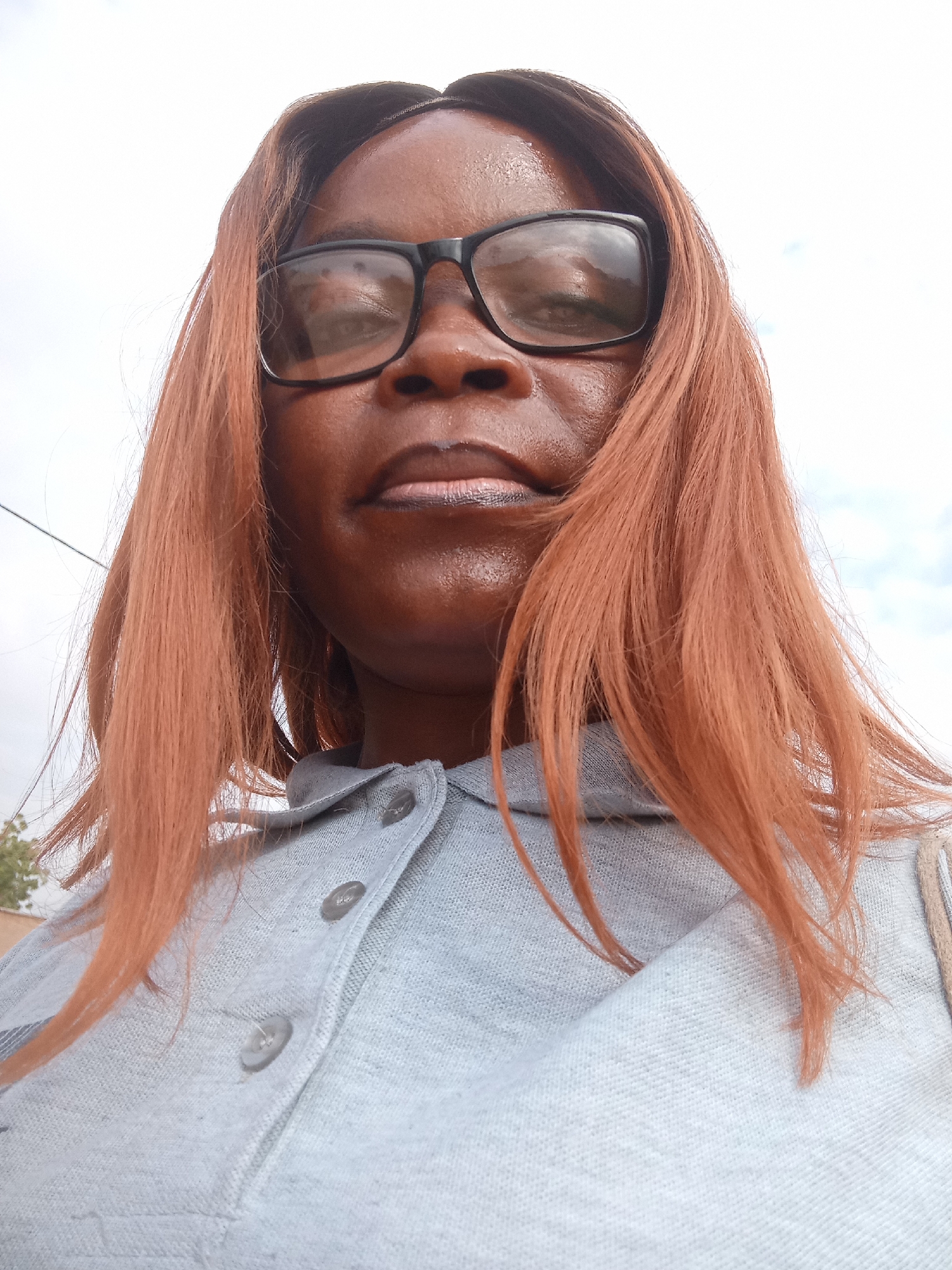 A low-angle, medium shot captures a Black woman from the chest up, her head tilted back slightly, looking upwards towards the camera. She has medium-toned brown skin, shoulder-length ginger-colored hair, and wears black-rimmed eyeglasses. Her mouth is closed, and her lips are painted with a metallic dark pink lipstick. She is wearing a light blue button-up collared shirt, with two visible grey buttons. The background is a pale, overcast sky with some light clouds. The image appears to have been taken outdoors in the city of Kason, Nigeria.