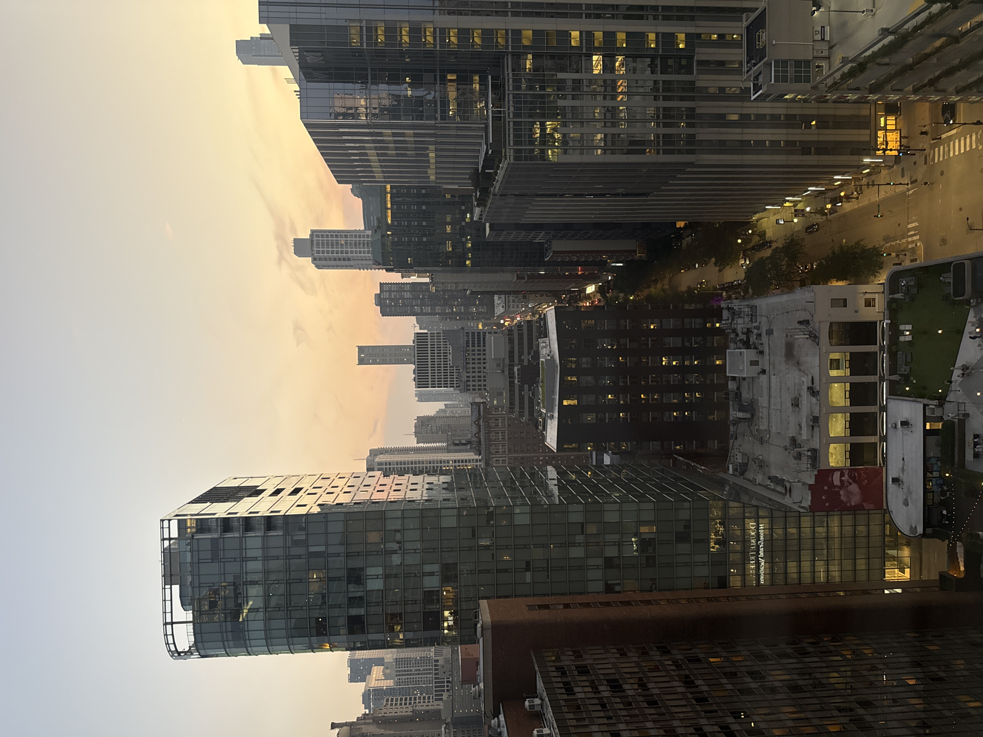 This is an aerial view of a city street in Chicago, Illinois, USA, at dusk. The scene is dominated by tall buildings with modern glass facades, interspersed with older brick structures. The sky is a soft gradient of orange, yellow, and pale blue, indicating the setting sun. Streetlights and illuminated windows create a warm glow in the urban landscape. Cars and pedestrians are visible on the street below, suggesting a bustling city environment. The perspective is from a high vantage point, looking down and across a cityscape.