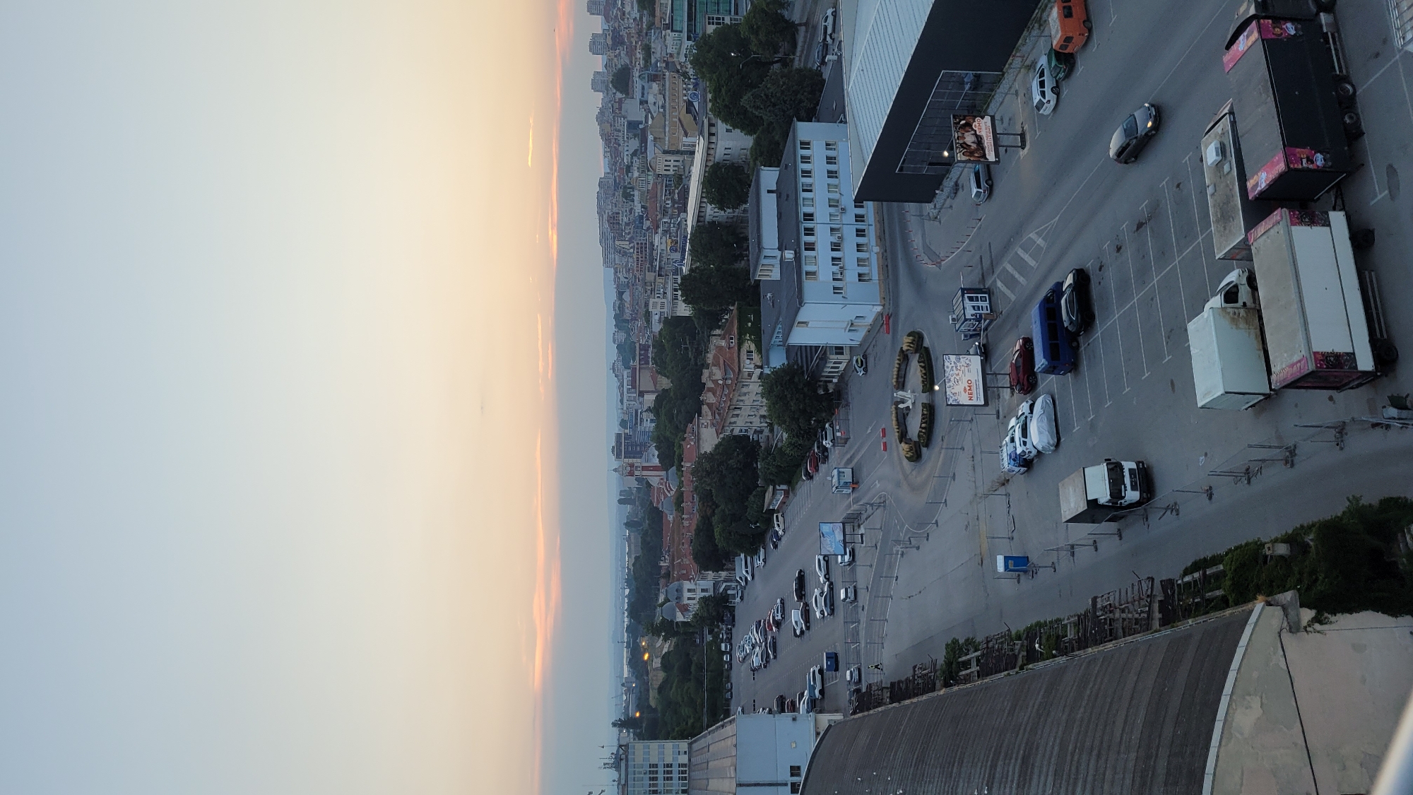 This is an aerial, wide-angle photograph taken at dusk in Varna, Bulgaria. The sun is setting, casting a soft, warm glow on the horizon and illuminating the sky with gradients of orange and blue. The image captures a cityscape characterized by a mix of modern and older buildings, interspersed with lush green trees.
In the foreground, a large, dark-colored building with a striped facade is partially visible, suggesting it might be a commercial establishment or industrial structure. Adjacent to it is a paved area, likely a parking lot, where several vehicles are parked. These include a white truck with its cargo bed open, a few cars, and several large trucks, some with trailers. One trailer is black and white, and another is white with pink and black graphics.
Further into the cityscape, a circular road or roundabout is visible, featuring decorative elements that appear to be animal statues, possibly lions, encircling a sign. Several cars are parked around this area. Beyond the immediate surroundings, the dense urban landscape of Varna stretches towards the horizon, with buildings of varying heights and architectural styles. The distant view hints at a body of water, possibly the Black Sea, given the location.
The overall scene depicts a quiet evening in a European city, with the day's activities winding down. The lighting suggests the transition from day to night, with the sky still holding onto the last rays of sunlight. No people are clearly discernible in the image. Visible text includes "OPEN" and "NEMO" on a sign near the roundabout.