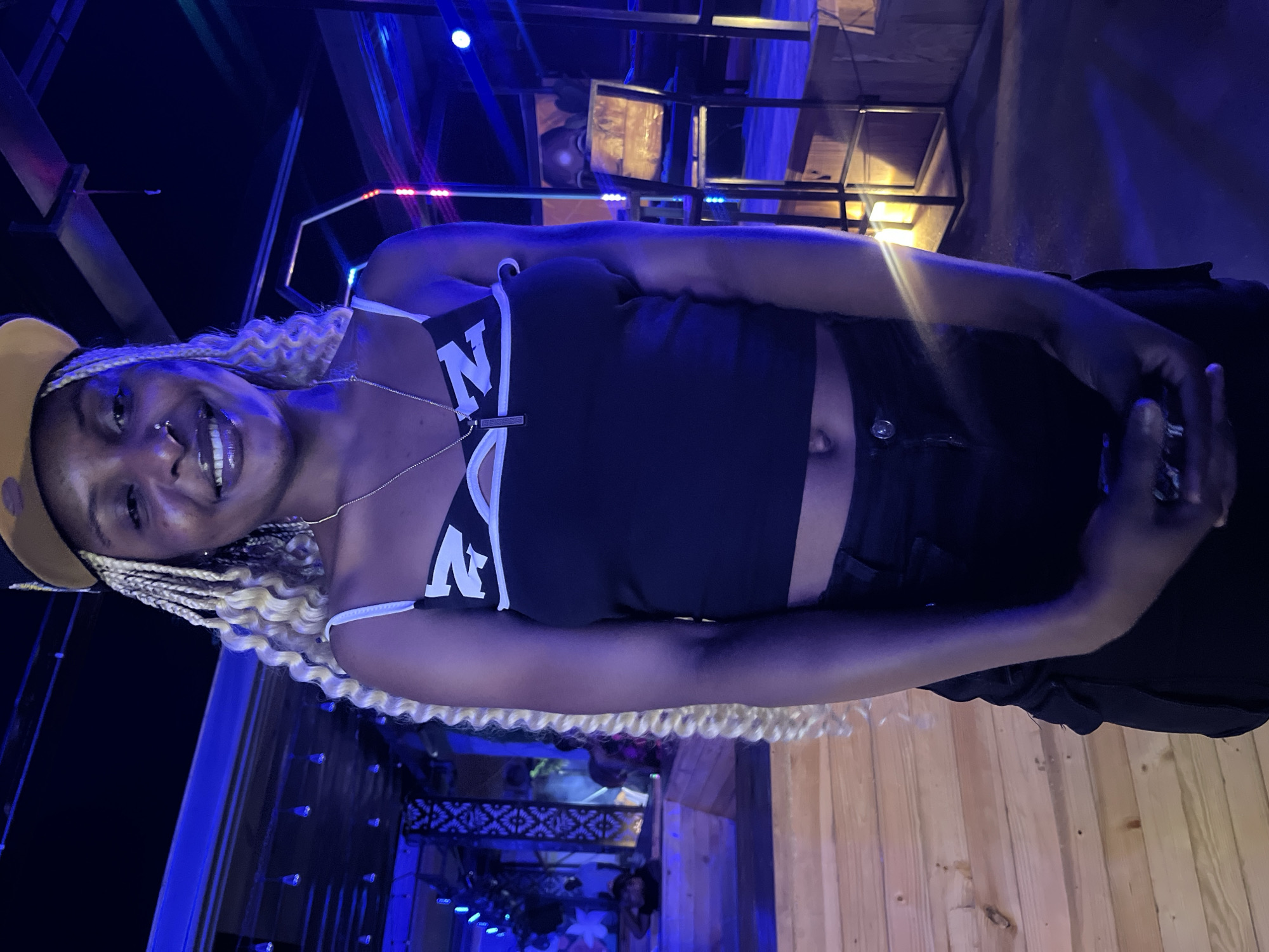 A woman is standing in a dimly lit indoor venue. She is wearing a black crop top with white trim and a black cap. Her hair is in blonde braids. She is smiling and holding a cell phone in her hands. The background is a blur of blue and purple lights, with wooden steps and railings visible. The environment appears to be a nightclub or bar, potentially located in Howayi, Nigeria.